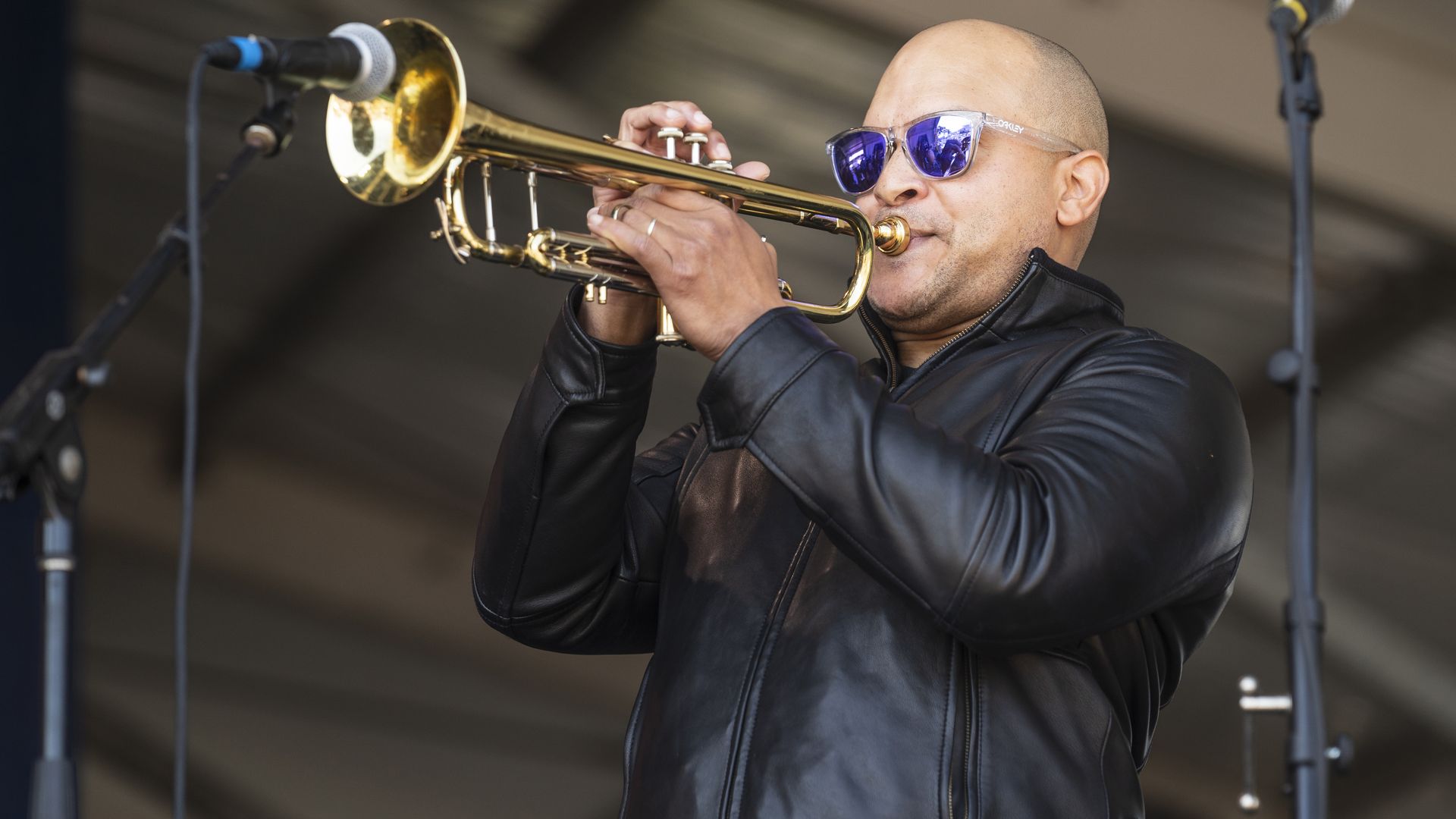 A man wears a leather jacket and purple-toned sunglasses while playing a trumpet onstage.