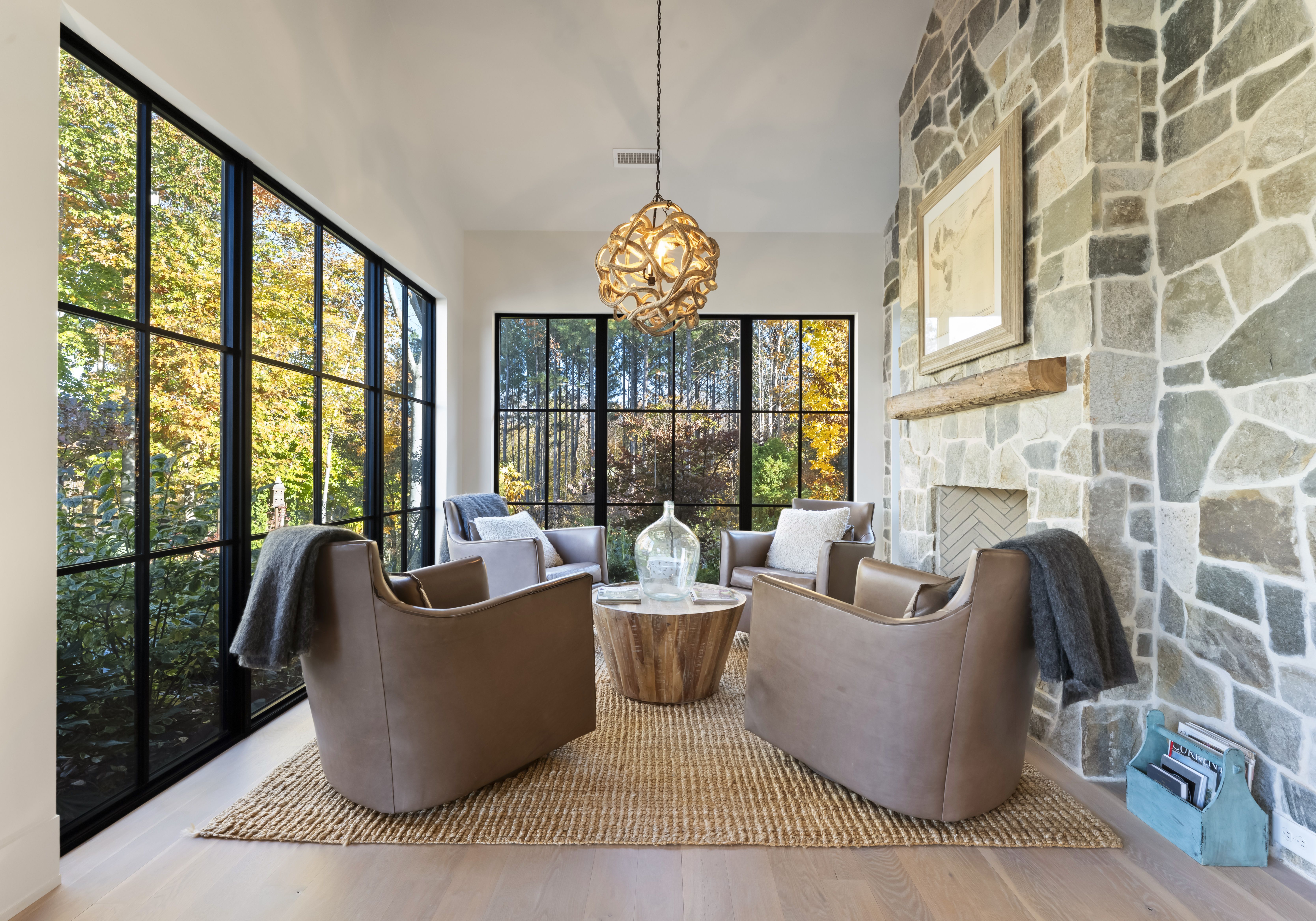 Cozy living room with large black-framed windows, a stone fireplace wall, a circular coffee table with a glass vase, four brown leather chairs, a pendant light, and a woven rug; autumn trees outside.
