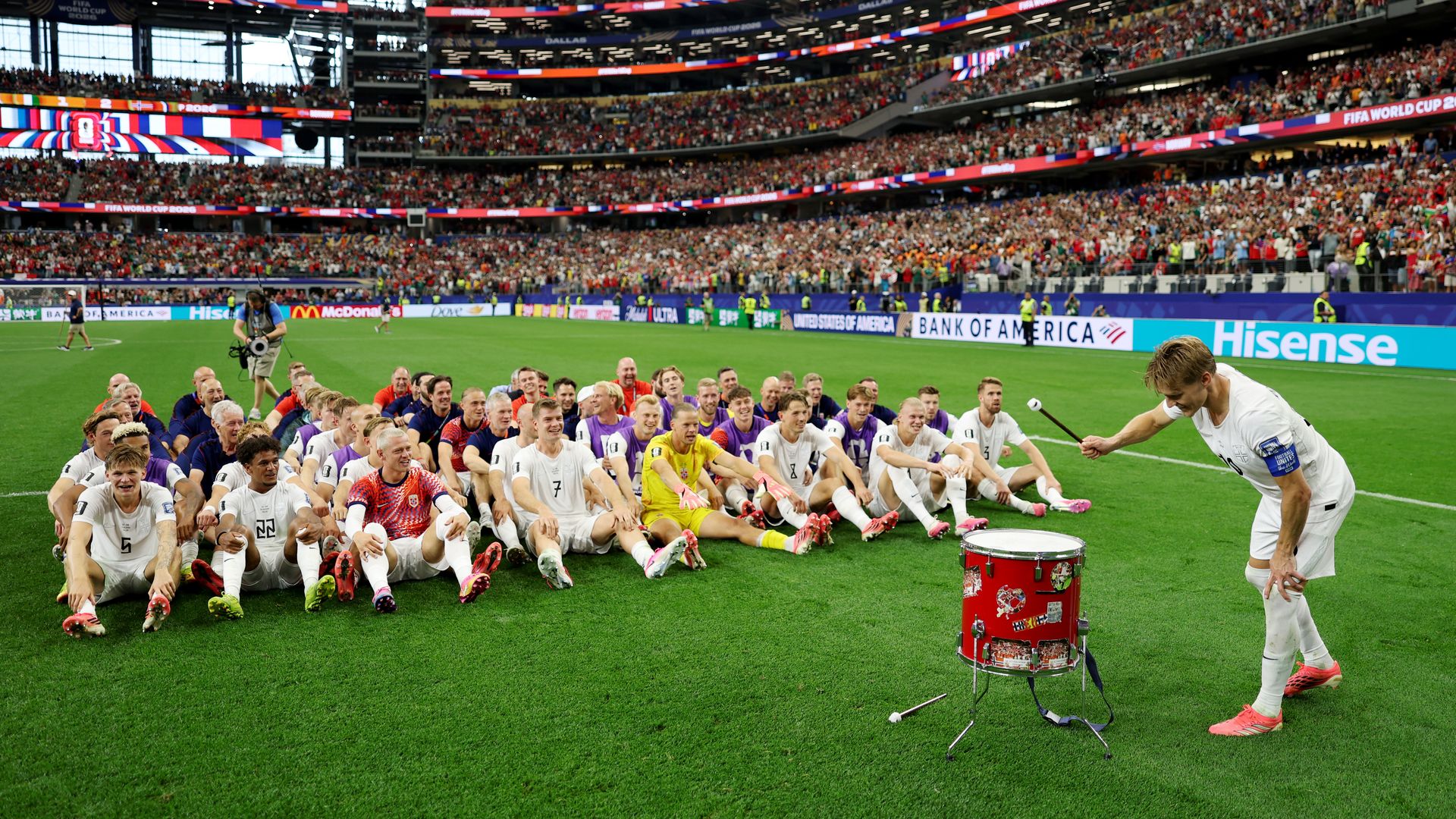 Norway national team player beating a drum with the rest of the team sitting in front of him on the pitch at Dallas Stadium after Norway's victory over Ivory Coast.