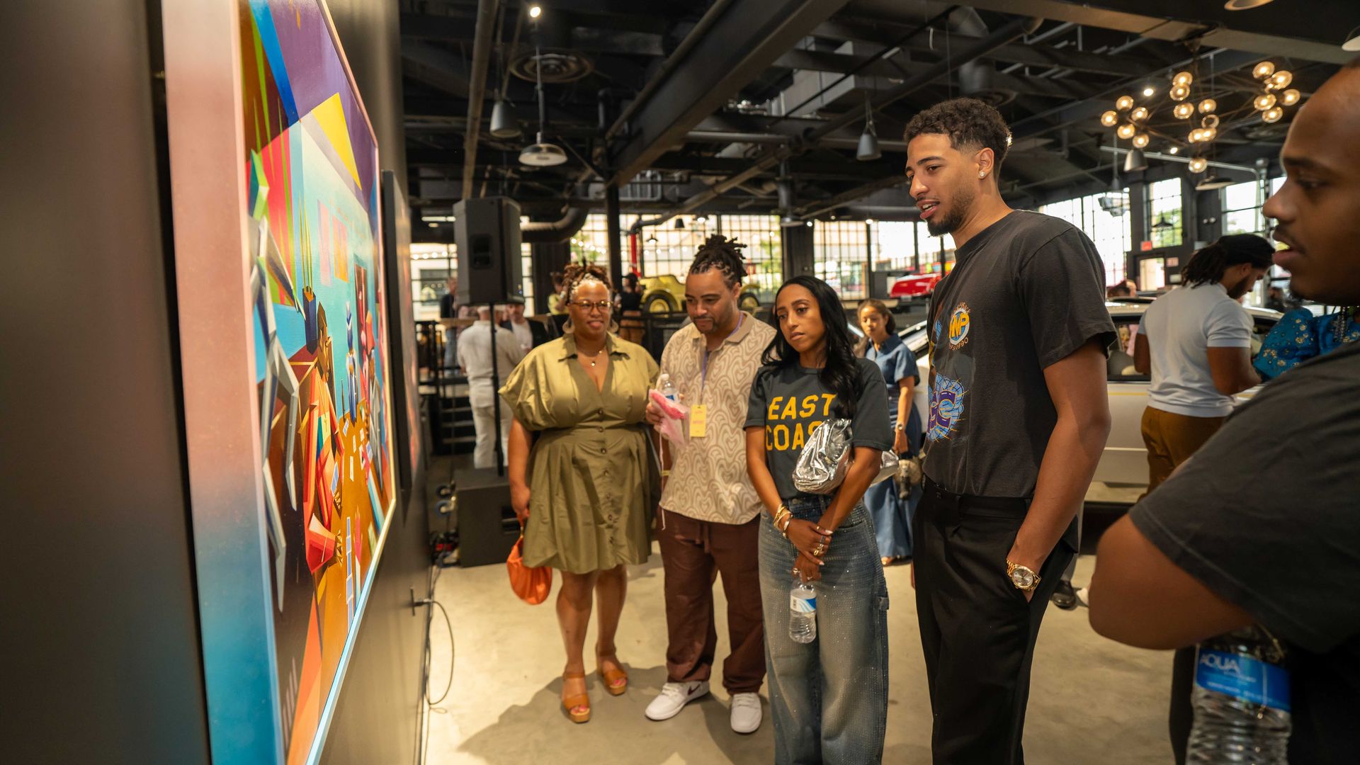 Group of people in casual clothing observing colorful modern artwork in a spacious gallery with industrial ceiling and large windows.