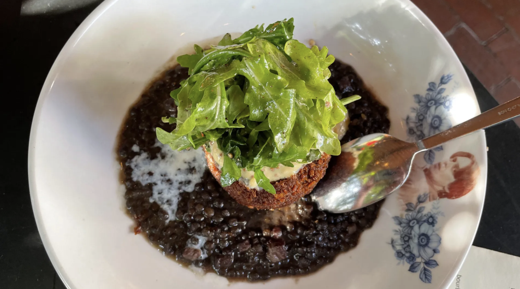 White bowl with black lentils, a fried patty topped with creamy sauce and fresh green arugula, accompanied by a silver spoon on a dark surface.