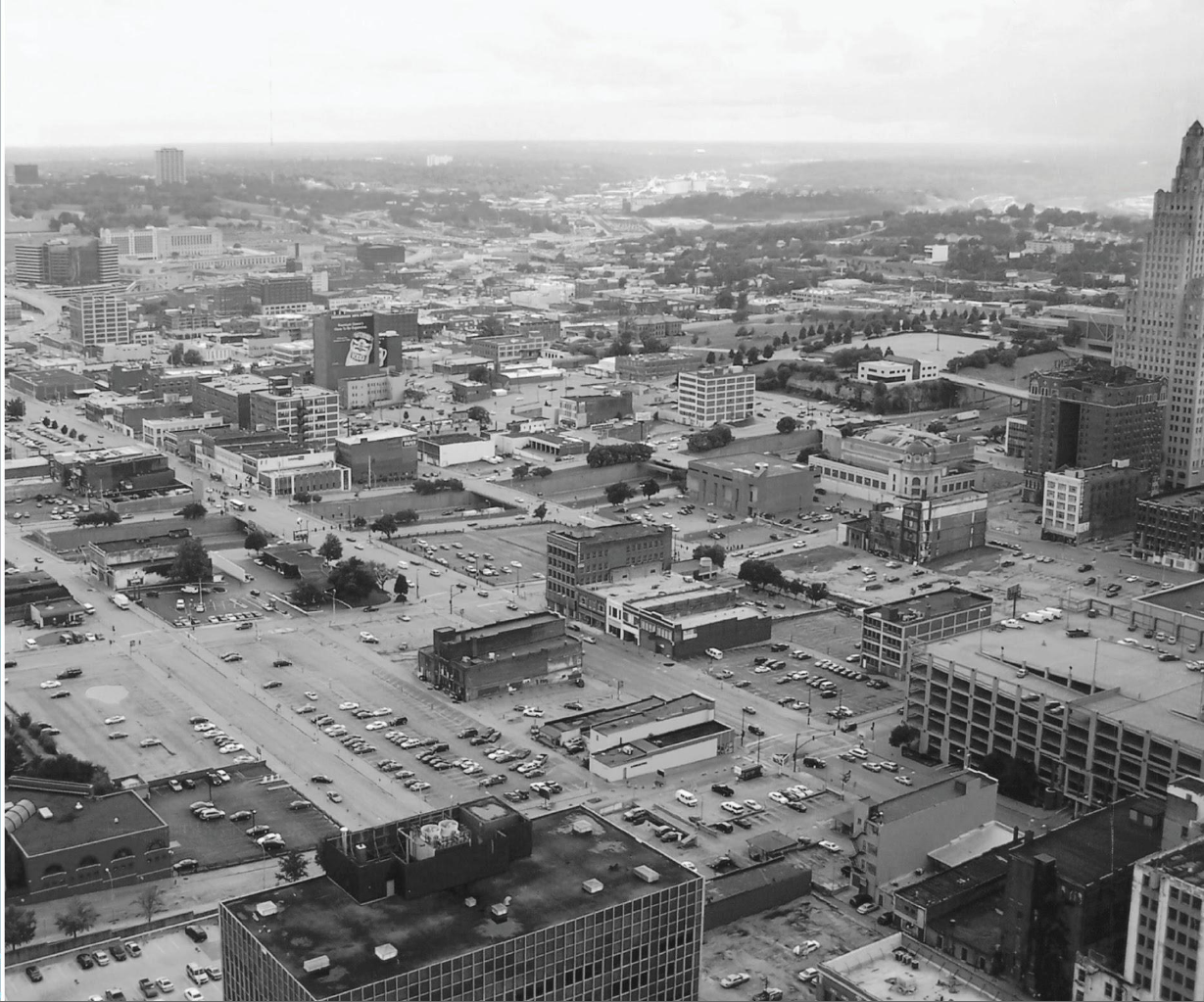 Black and white aerial view of a sprawling city with many low-rise buildings, parking lots filled with cars, and a tall skyscraper on the right side under a cloudy sky.