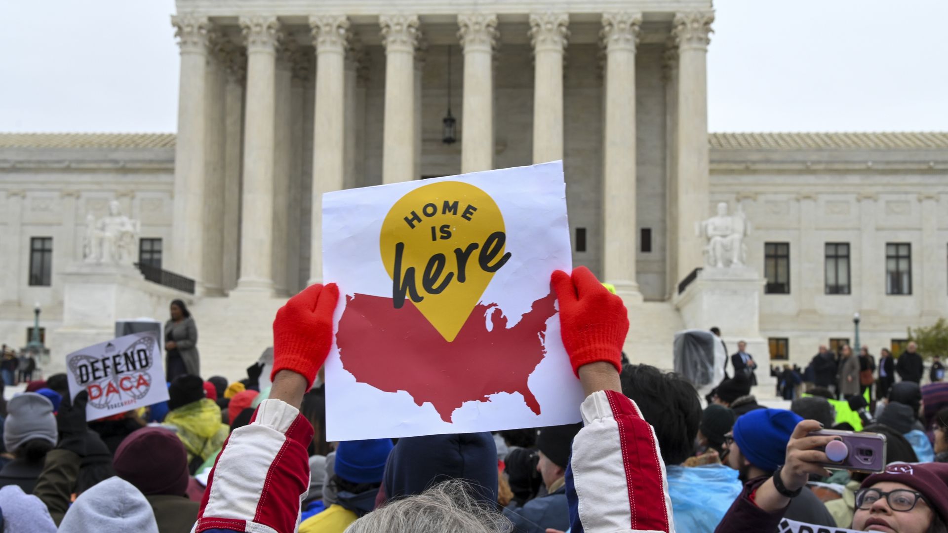 Protestors in front of the Supreme Court. One protestor in the center holds up a sign that says "home is here." 