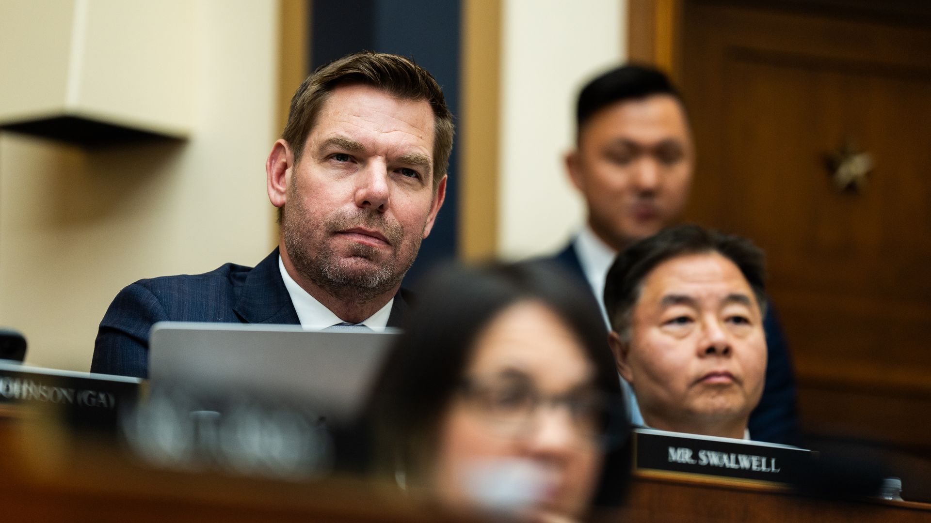 A man in a navy suit and white shirt sits at a formal hearing desk with a laptop, looking toward the camera; blurred colleagues and a nameplate reading "MR. SWALWELL" appear in the background.