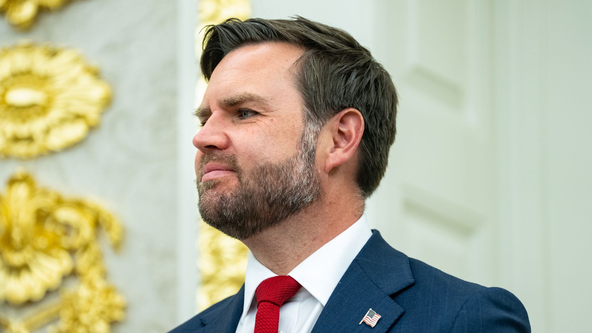 JD Vance, wearing a red tie and an American flag pin on his lapel, looks on in the Oval Office.