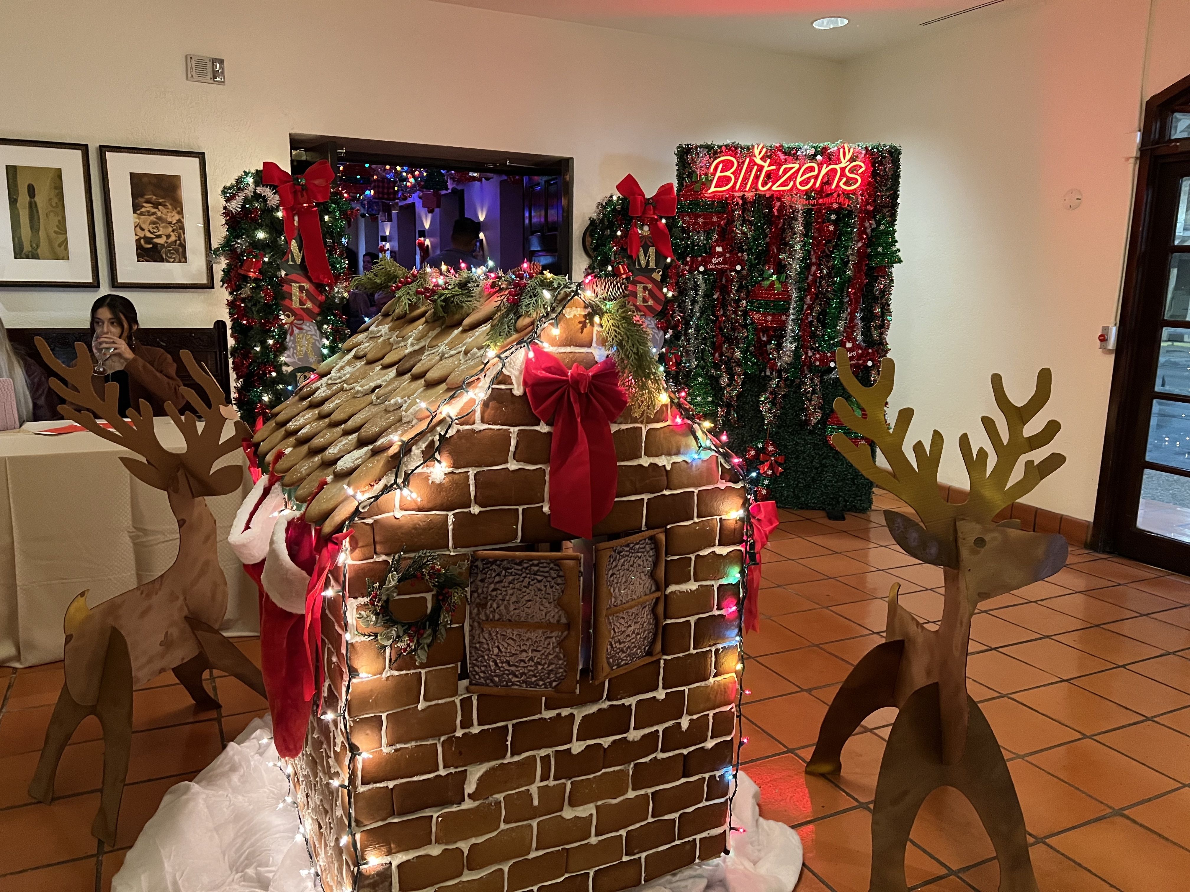 Gingerbread house decorated with lights, red bows, and greenery, flanked by two wooden reindeer; festive Christmas wreaths and a red neon sign reading "Blitzen's" in the background indoors.