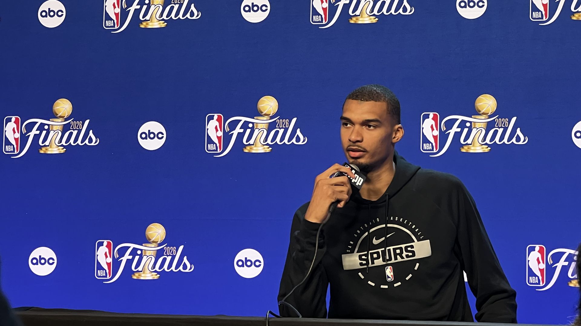 Blue backdrop with repeated NBA Finals logos and ABC circles; a man in a black Spurs hoodie speaks into a microphone at a press conference.
