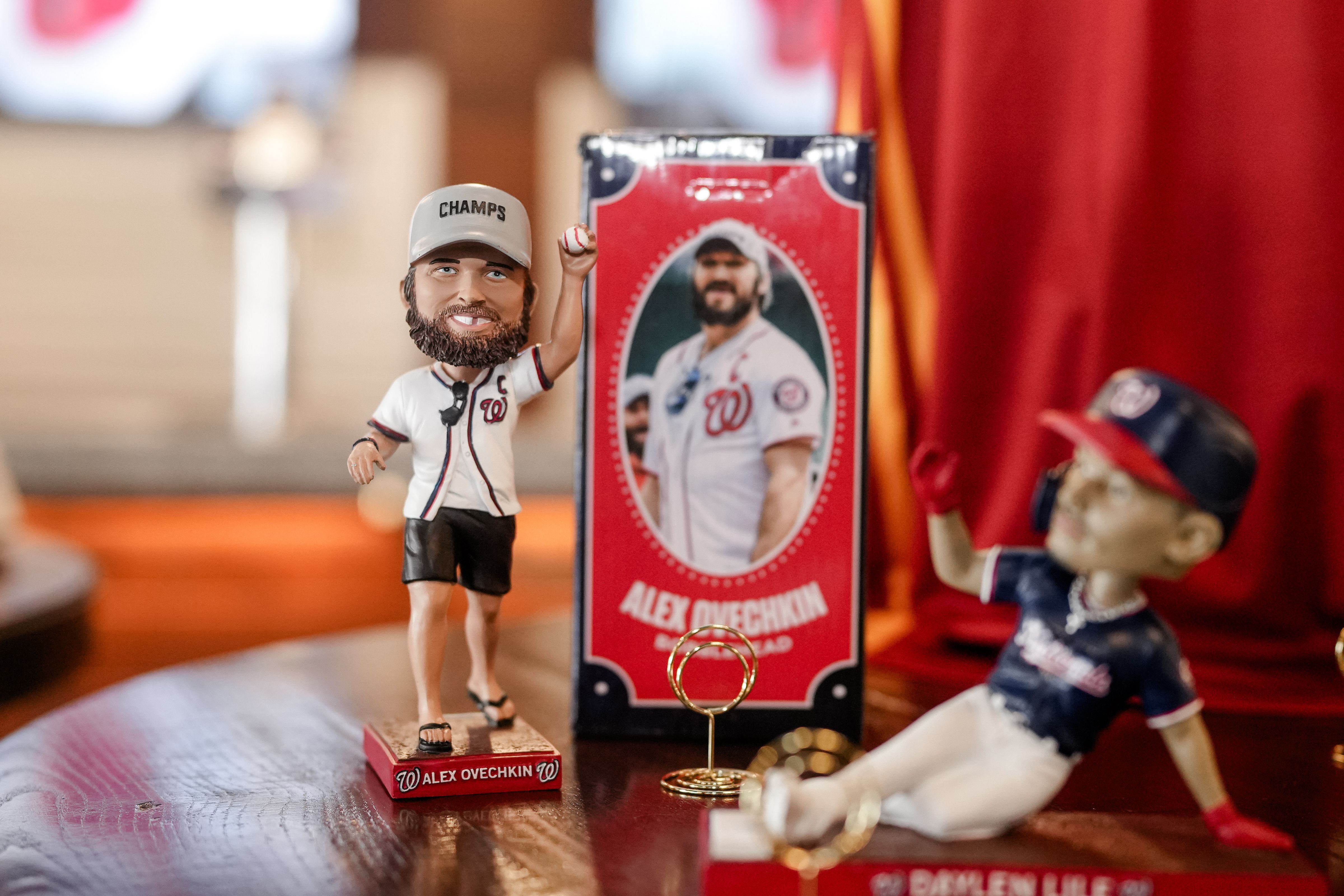 Bearded bobblehead of Alex Ovechkin in a white Nationals-style jersey and a "CHAMPS" cap stands on a red base beside a red "Alex Ovechkin" box; a blue-uniform figure sits on the right on a wooden table.