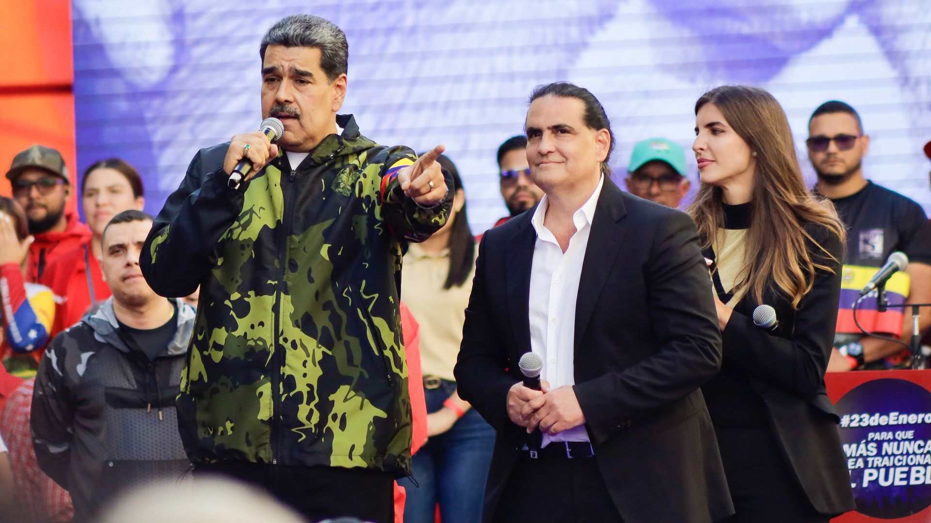 Man in camouflage jacket speaks into a handheld microphone and points, flanked by a suited man and a woman on a stage with a blue backdrop and a cheering crowd.