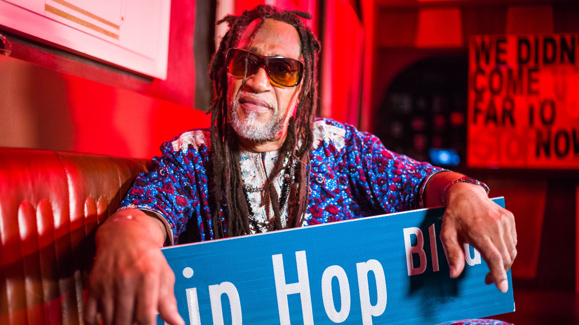 Photo shows DJ Kool Herc, the father of hip-hop, sitting on a leather couch and holding a sign that says "Hip Hop Blvd."