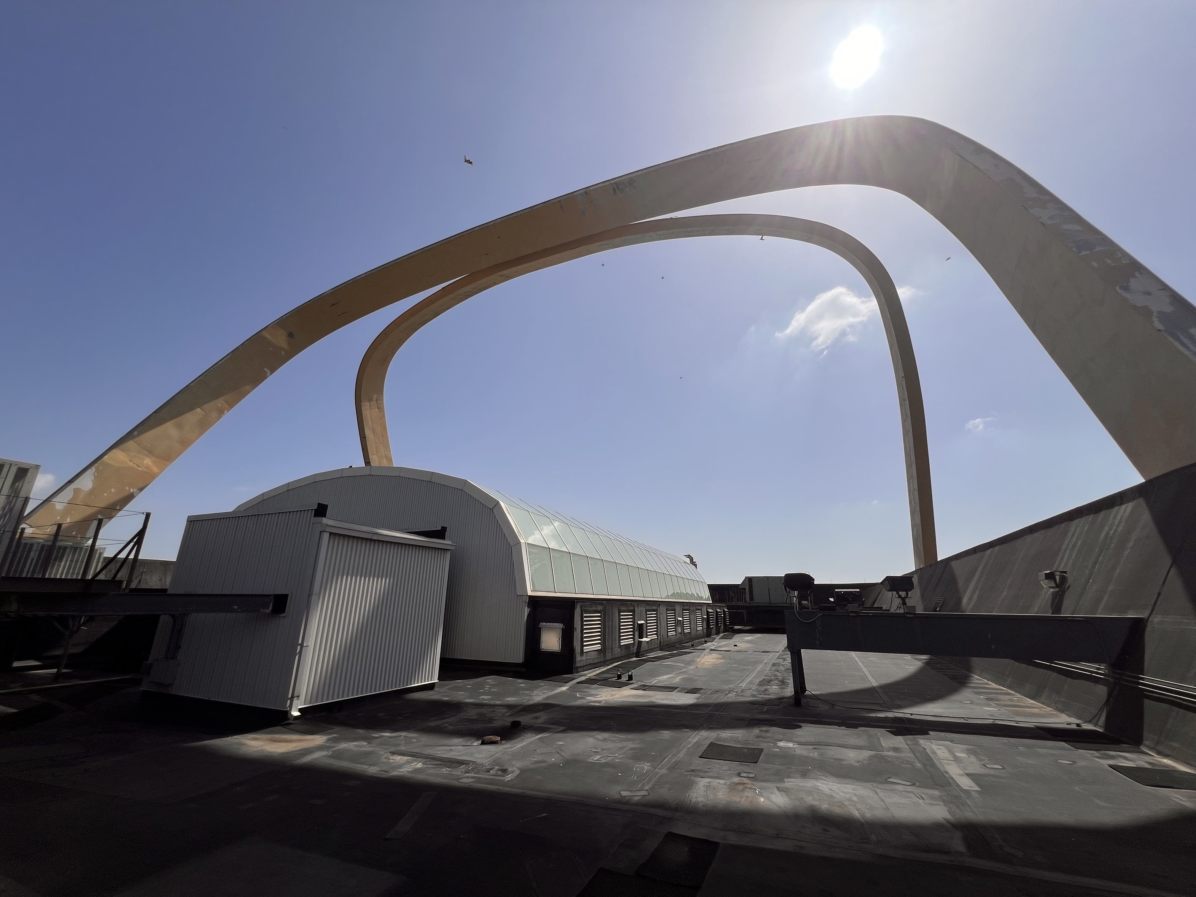 Clear blue sky over a rooftop with massive curved beige metal arches; sun glare at the top right; industrial buildings along the lower edge; a small flying object in the distance.