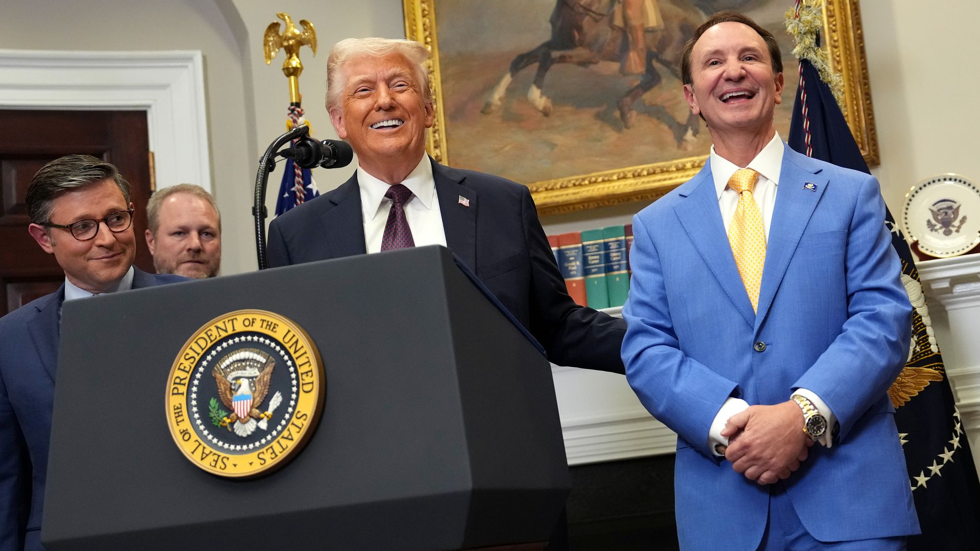 Standing at a podium with the presidential seal, President Donald Trump smiles and puts an arm around the back of Gov. Jeff Landry, who stands to Trump's left. Speaker Mike Johnson looks on smiling from Trump's right.