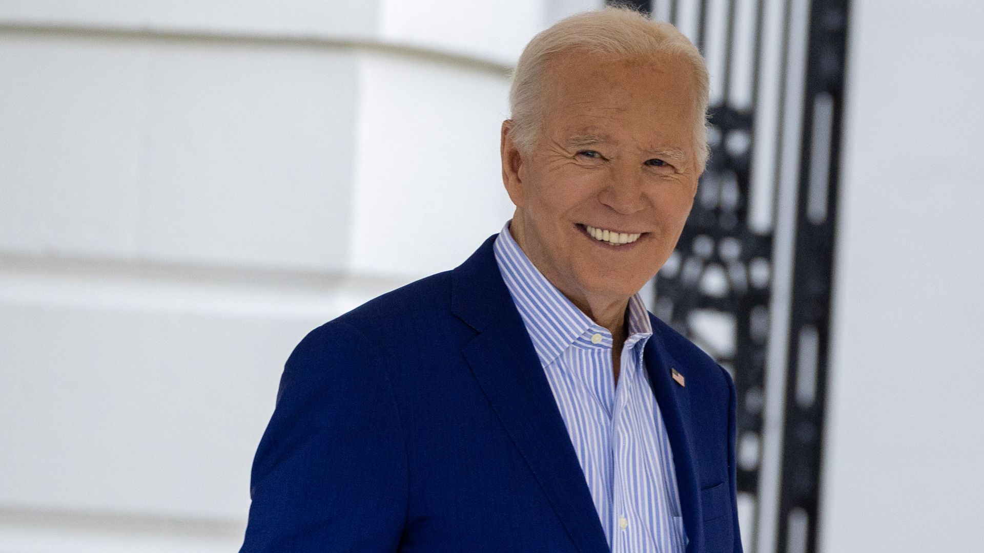 Joe Biden, wearing a blue jacket and a blue and white stripped shirt, smiles as he leaves the White House.
