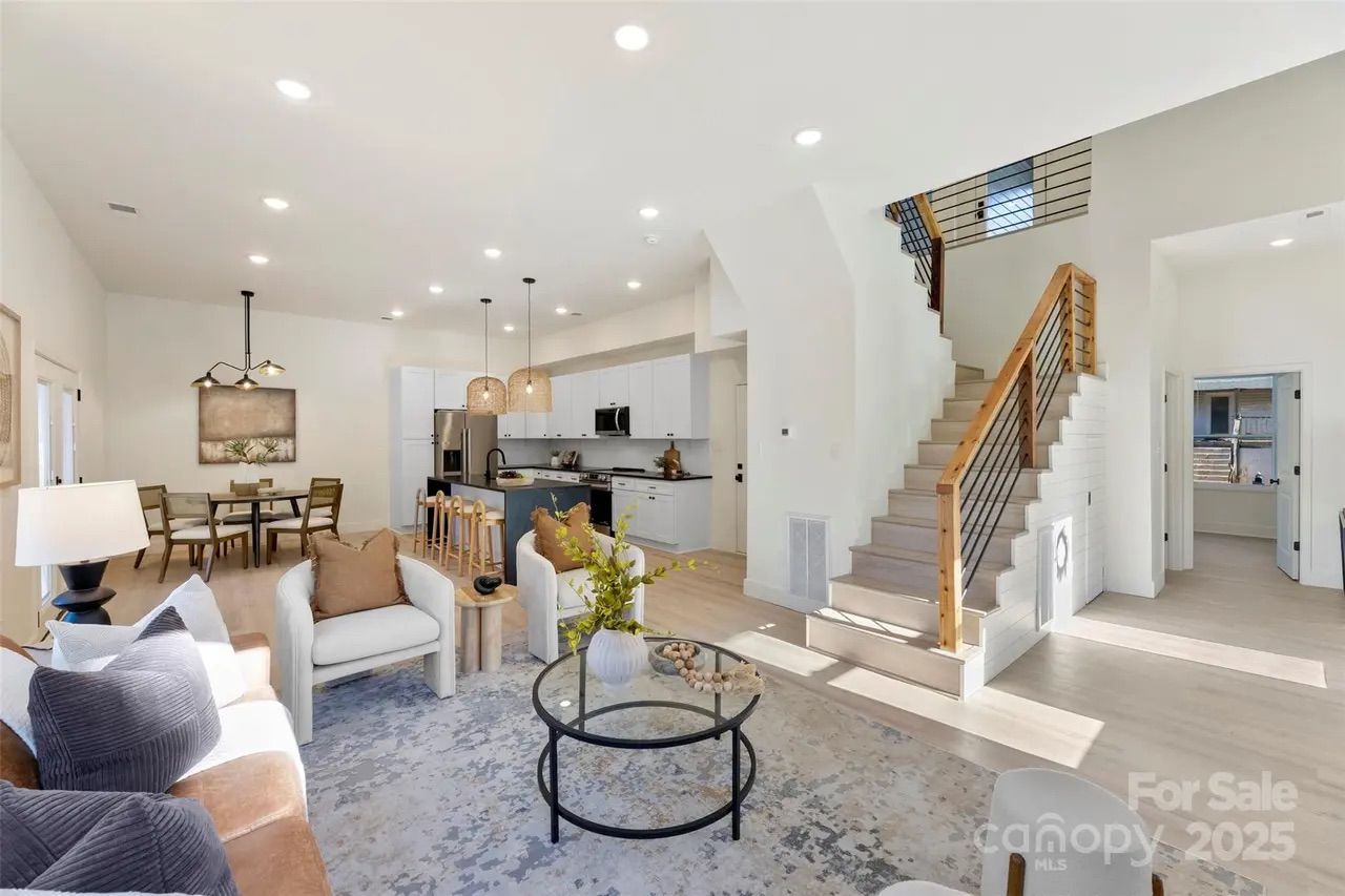Bright open living room with beige sofa, white armchairs, glass coffee table, light wooden floor, staircase with wooden railing, white walls, dining table, and modern kitchen with hanging lights.