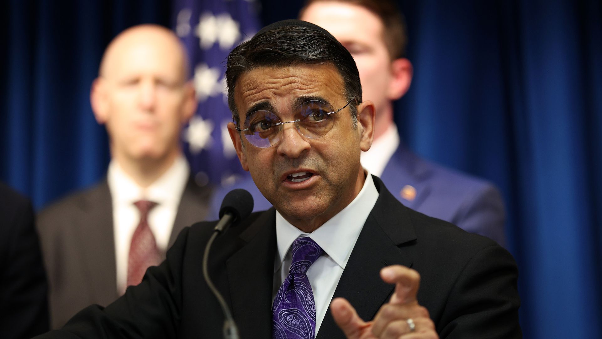 Daniel Rosen in a dark suit and purple tie speaks at a microphone, wearing glasses. Behind him are blurred figures and a blue backdrop with part of the American flag visible.
