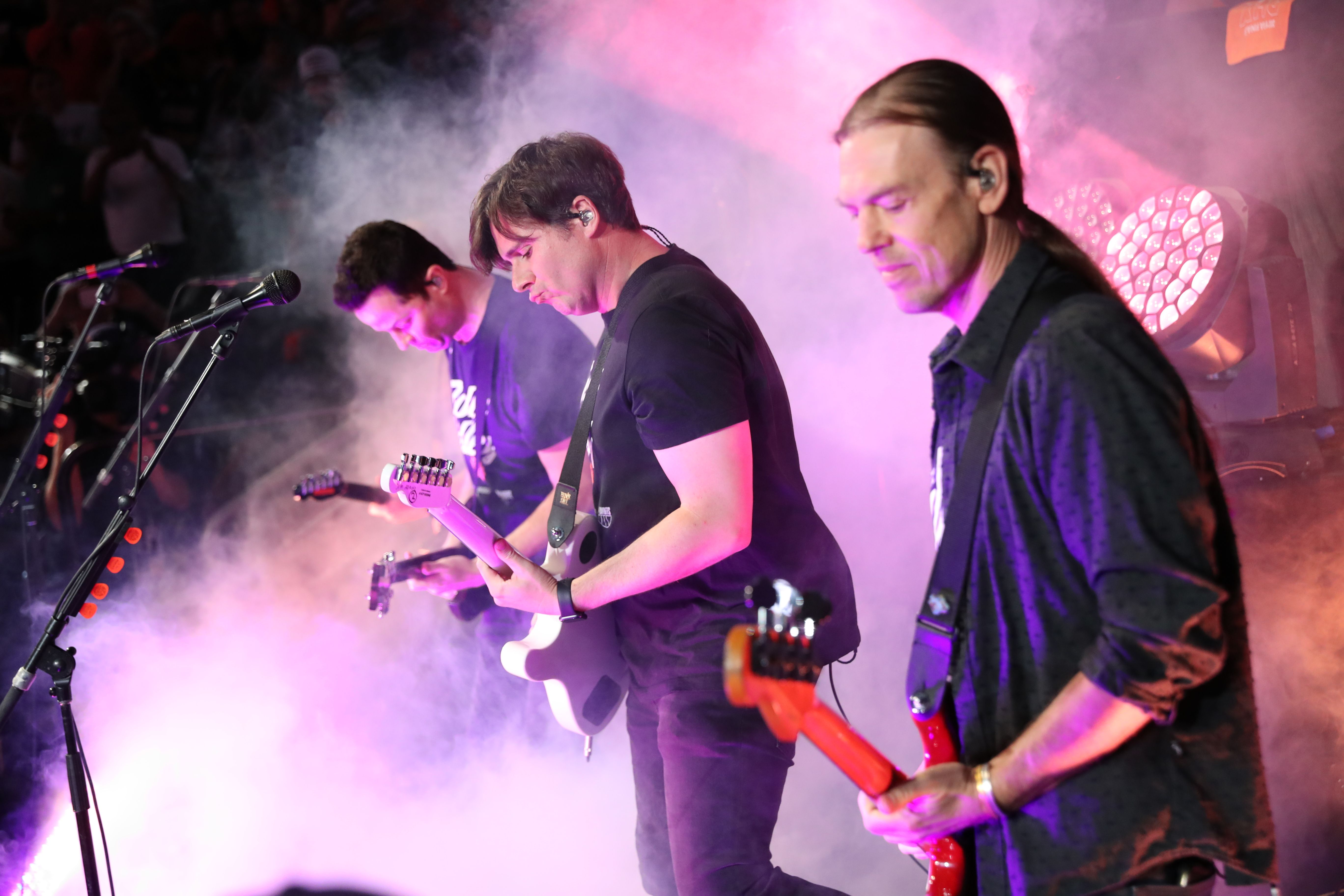three men standing on stage with electric guitars, with purple hues and fog surrounding them. 