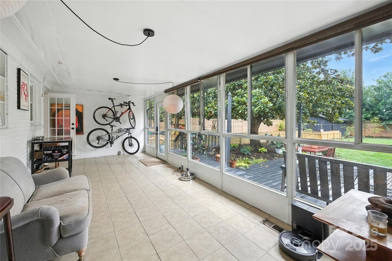 Bright sunroom with large windows overlooking a garden, beige tiled floor, gray sofa, two bikes mounted on white wall, hanging paper lantern, and a small table with objects.
