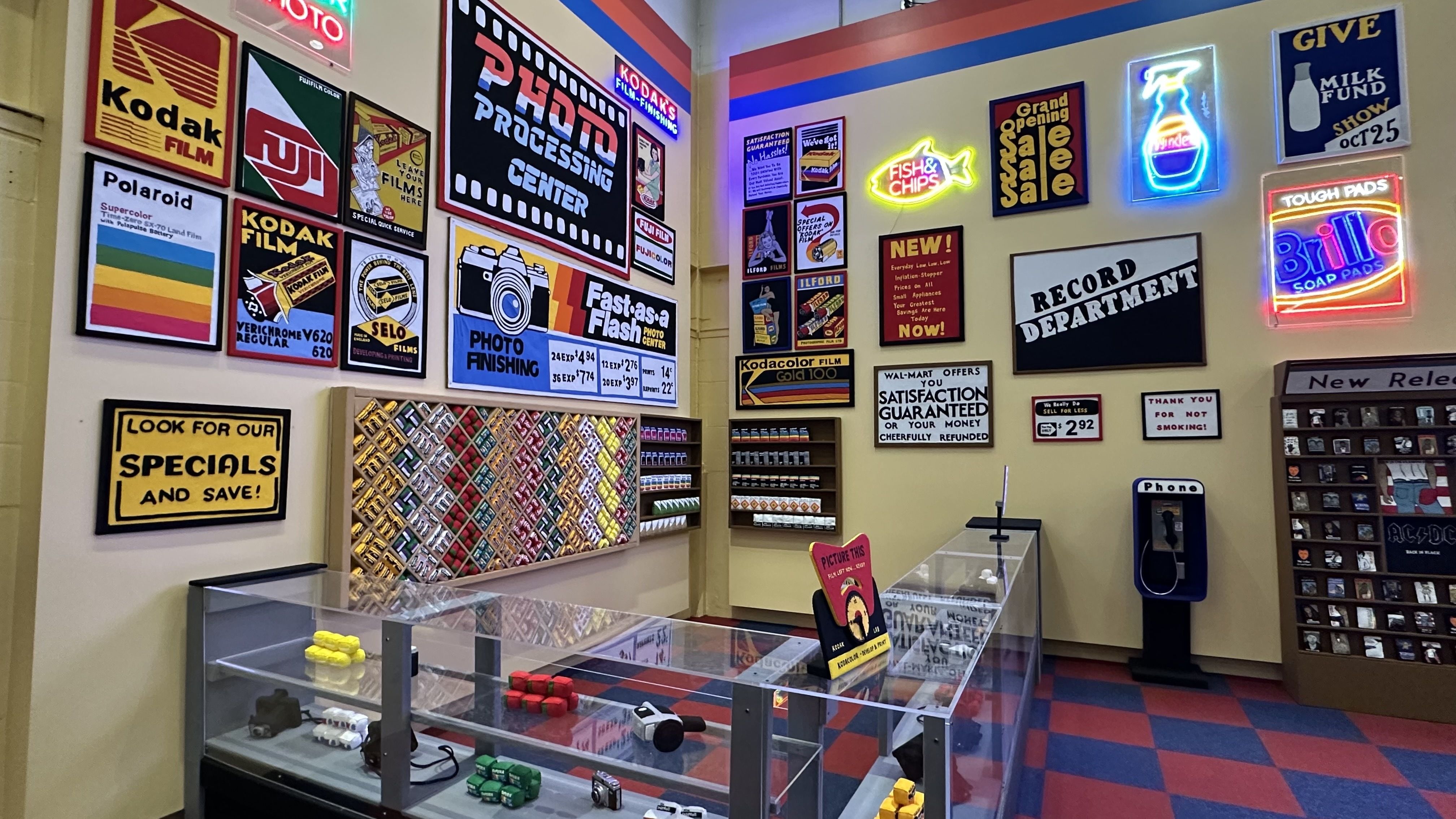 Retro photography shop wall: cream walls covered with bright vintage posters and neon signs (Kodak, Polaroid, Fish & Chips); glass display cases and a blue-red checkered floor.