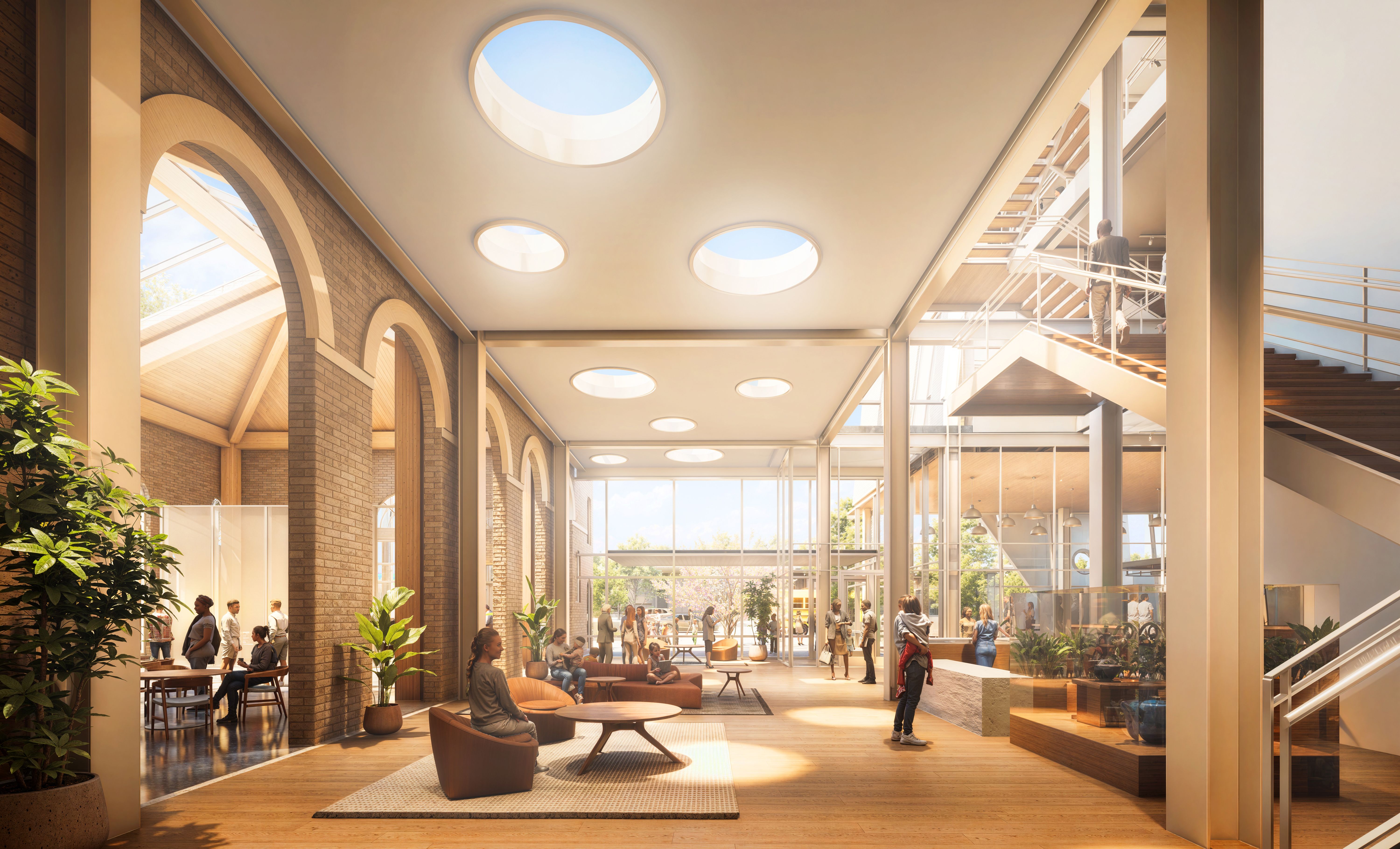 Bright, modern indoor space with wooden floors, brick arches, circular skylights, plants, and people sitting, walking, and talking, with a staircase on the right and large windows letting in daylight.