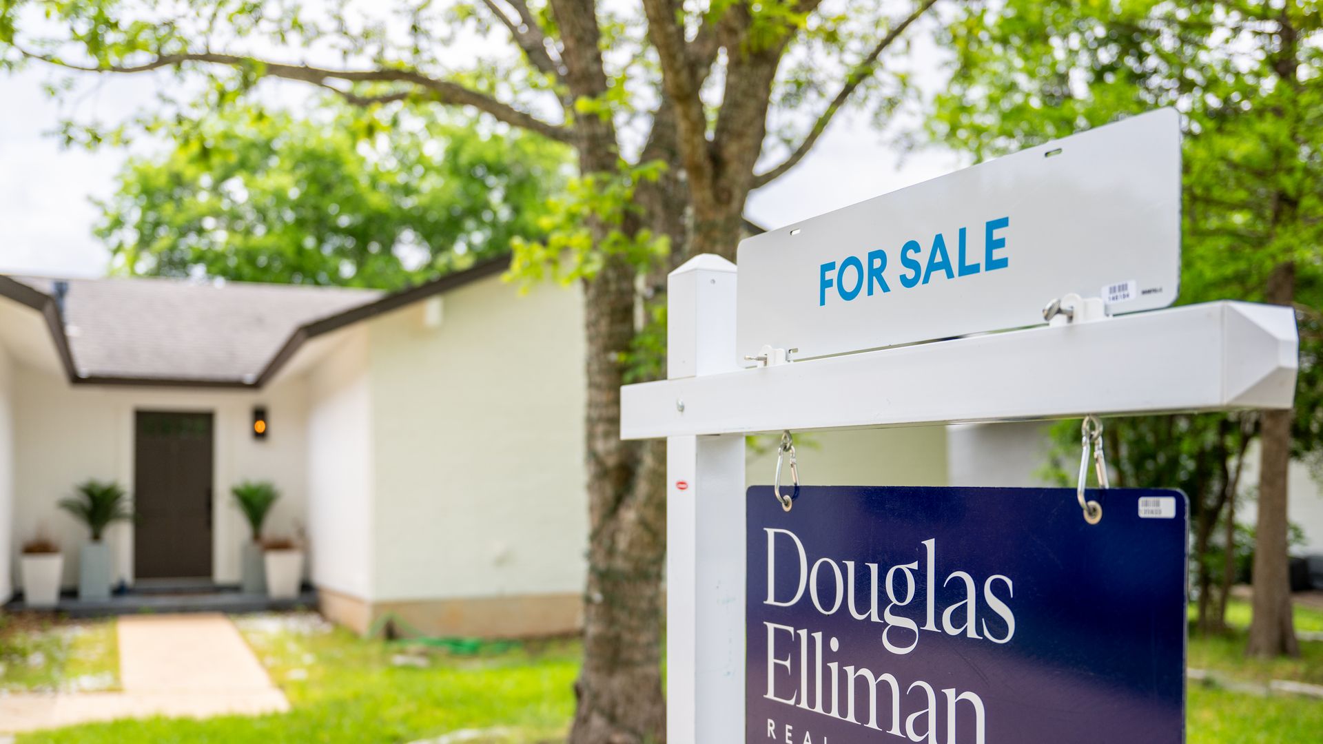 White house with a brown door and green lawn, featuring a white and blue "For Sale" sign from Douglas Elliman Real Estate in the foreground, with trees and greenery around.