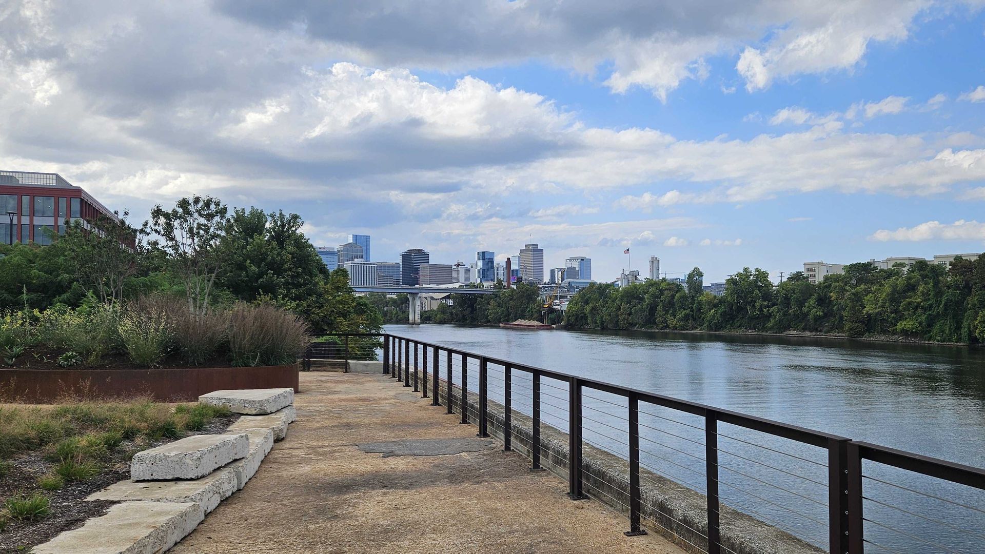 Riverside park path with a black railing along a calm river, leading to a distant city skyline and bridge. Lush trees line both banks under a blue sky with white clouds.