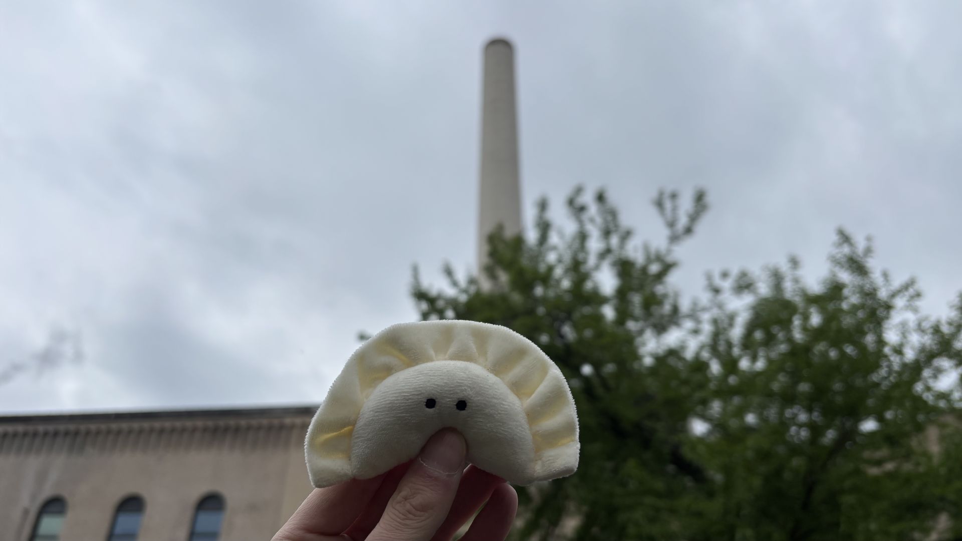 Hand holds a white plush dumpling with two black eyes in the foreground; behind it are a tall concrete smokestack, cloudy sky, and green trees by a brick building.
