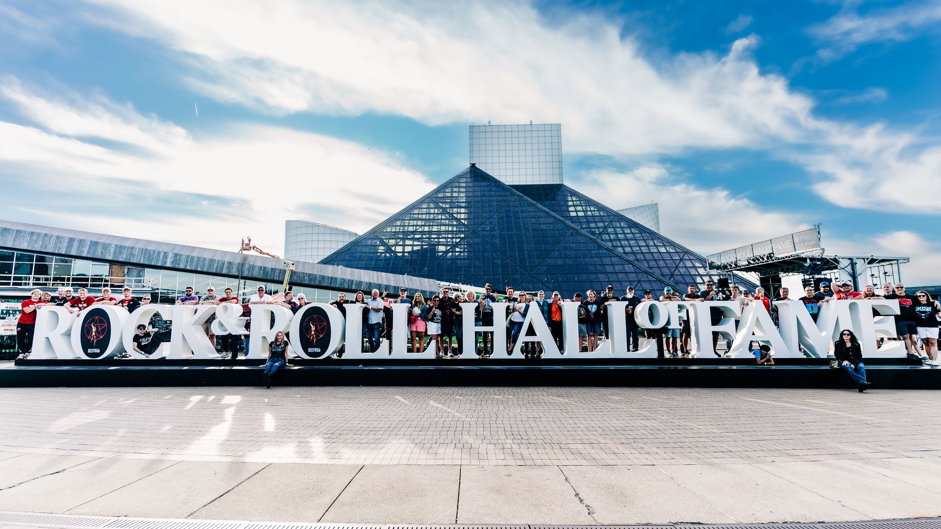 A large crowd stands behind oversized white letters reading "ROCK & ROLL HALL OF FAME" with a glass pyramid building and blue sky in the background.