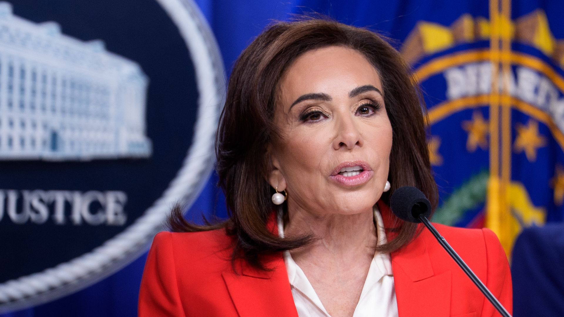 Portrait of a woman, wearing a red blazer, speaking into a microphone at a press conference; blue backdrop with government seals and the word "JUSTICE" is visible.