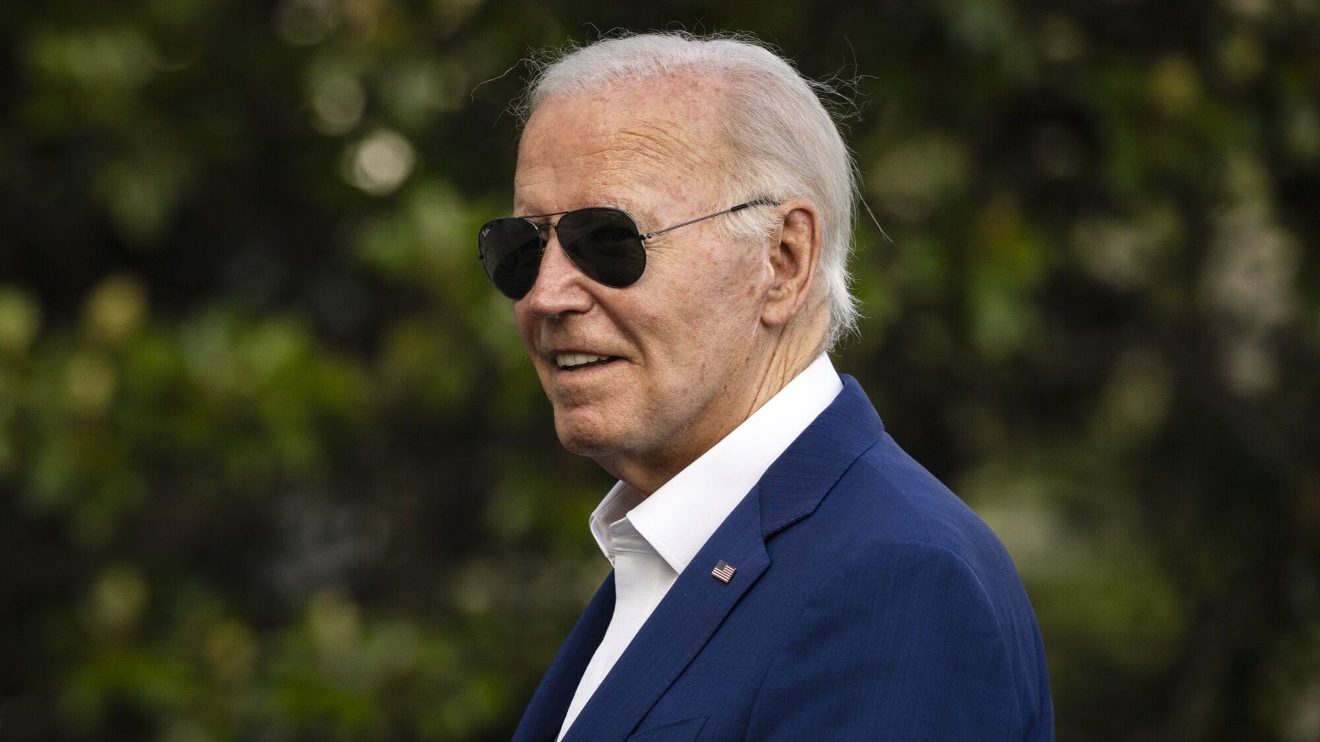 President Biden, wearing a blue suit and aviators standing in front of a tree.