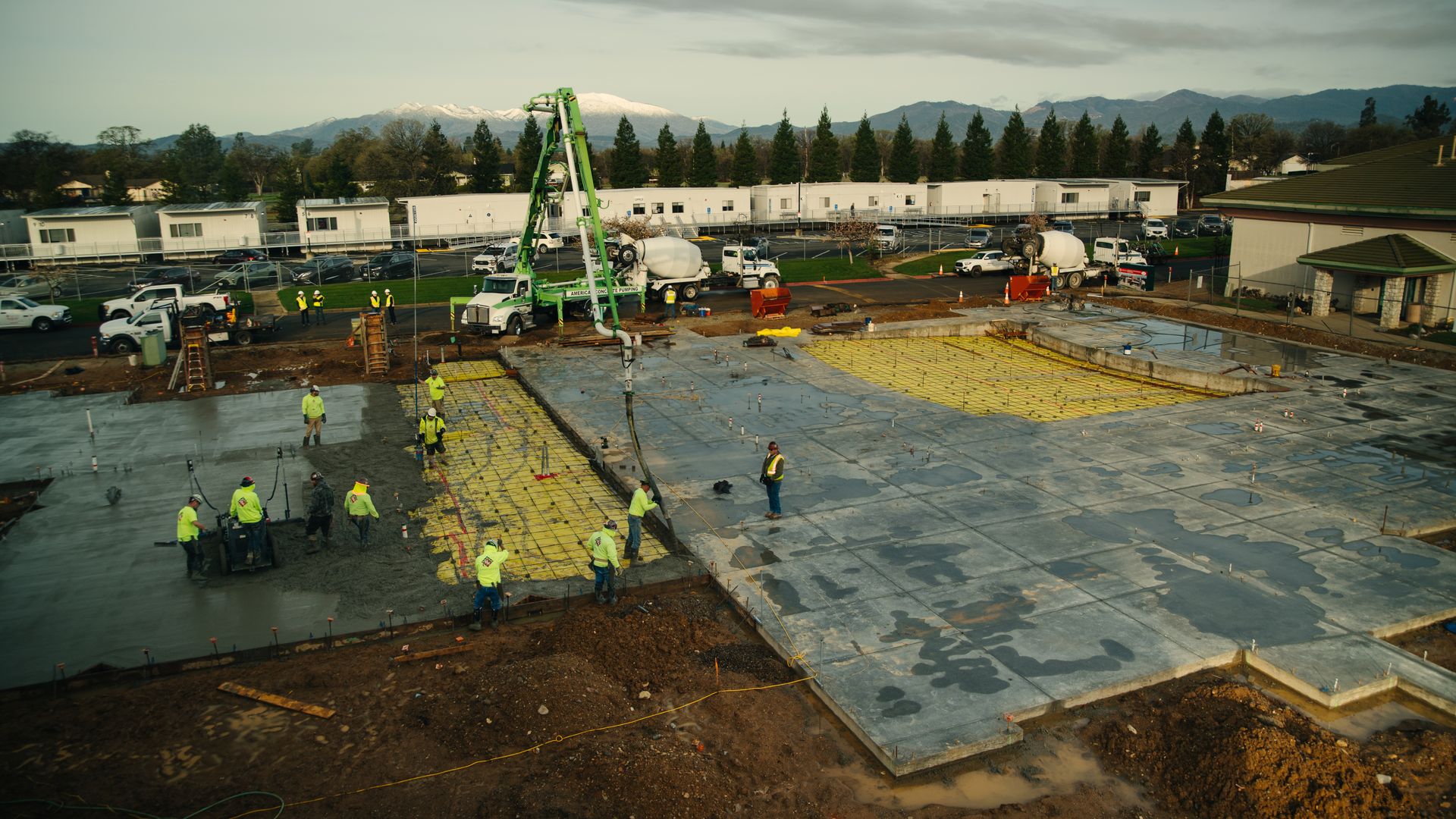 Construction workers in yellow safety vests pouring concrete on foundation slabs with rebar, trucks, and equipment around, with snow-capped mountains and trees in the background.