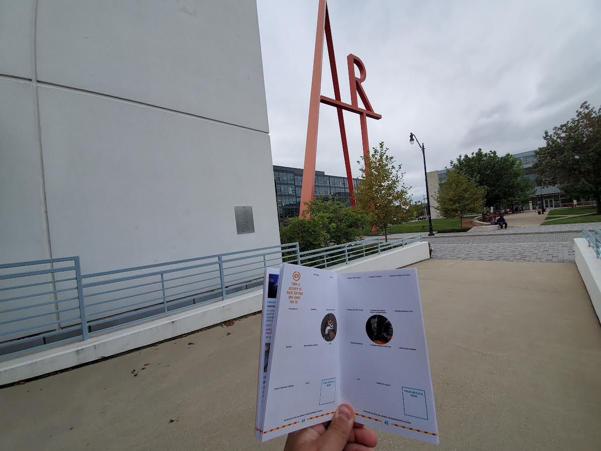 Holding up an "Art Passport" program brochure near an art college's large outdoor exhibit spelling the word "ART"