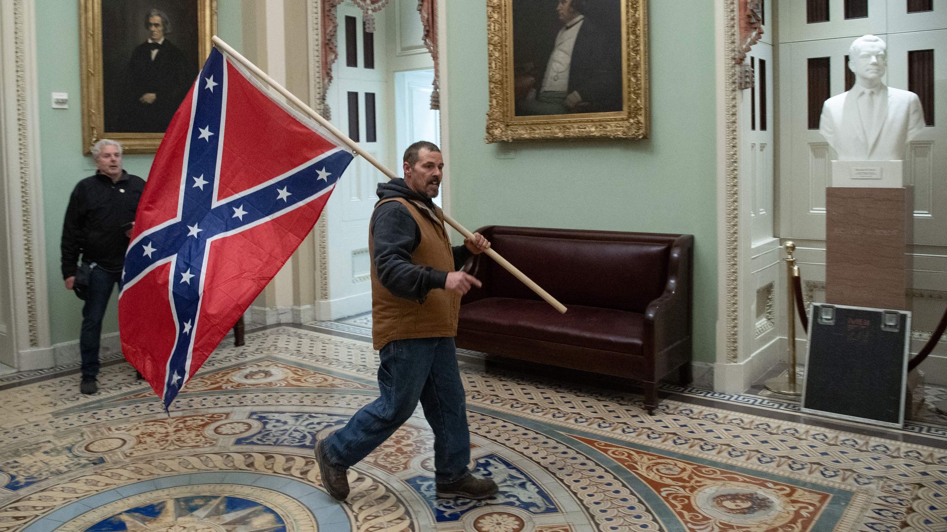 Photo of a man holding a Confederate flag as he walks in the Capitol building hallways