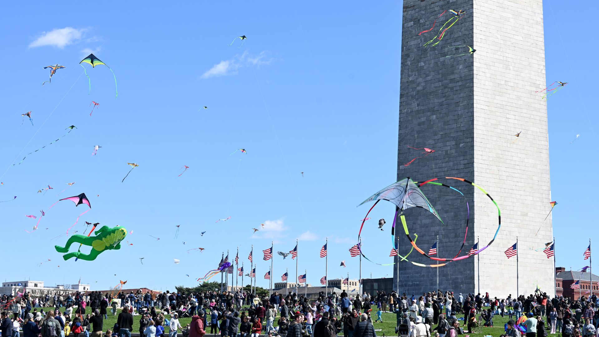 Kites are flying in the air in a picture taken on the grounds of the Washington Monument, visible in the background against a sunny, blue sky