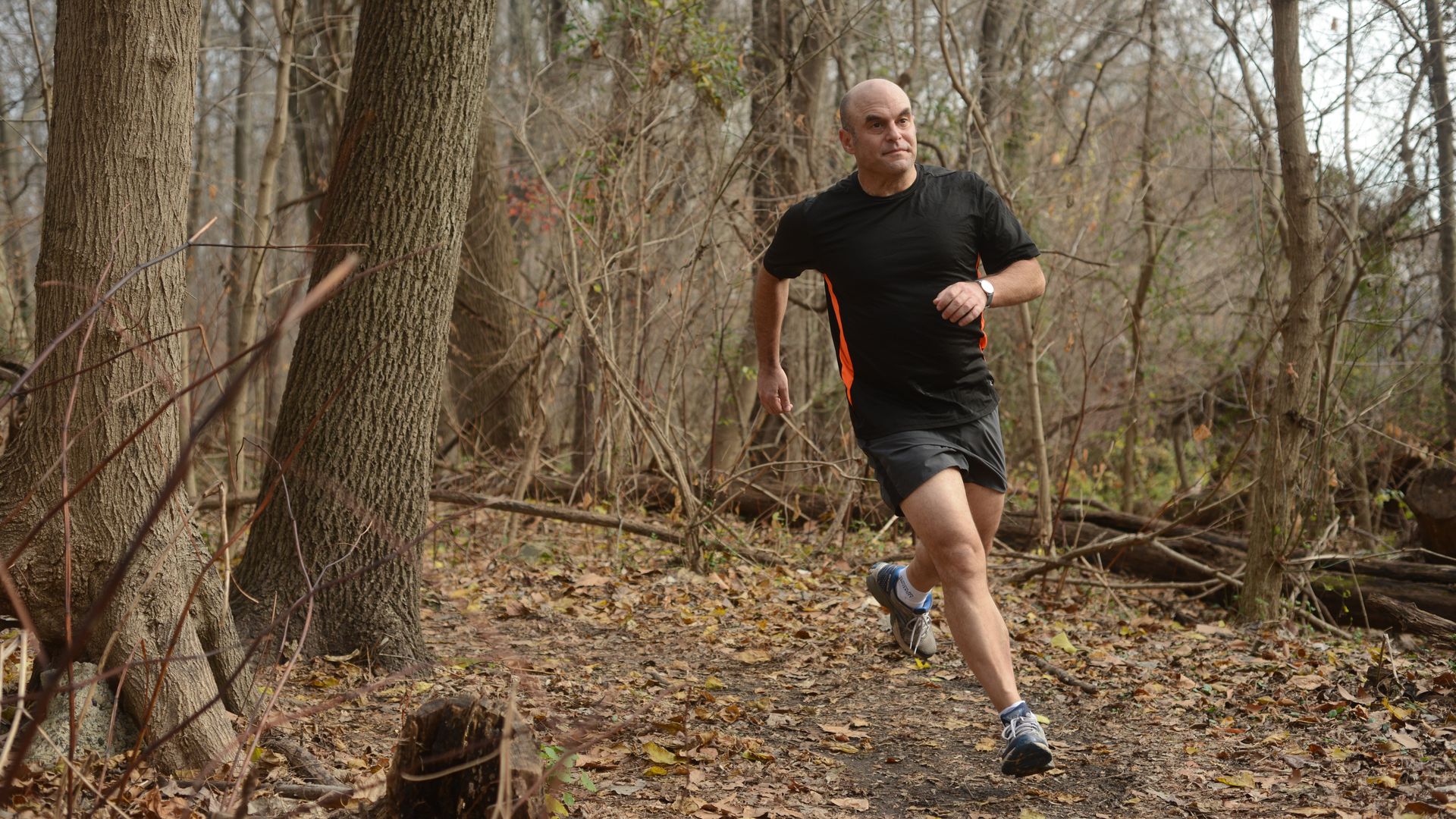 A man in black shorts and a black t-shirt runs around the bend of a trail in a forested park. The trees in the background have lost their leaves.