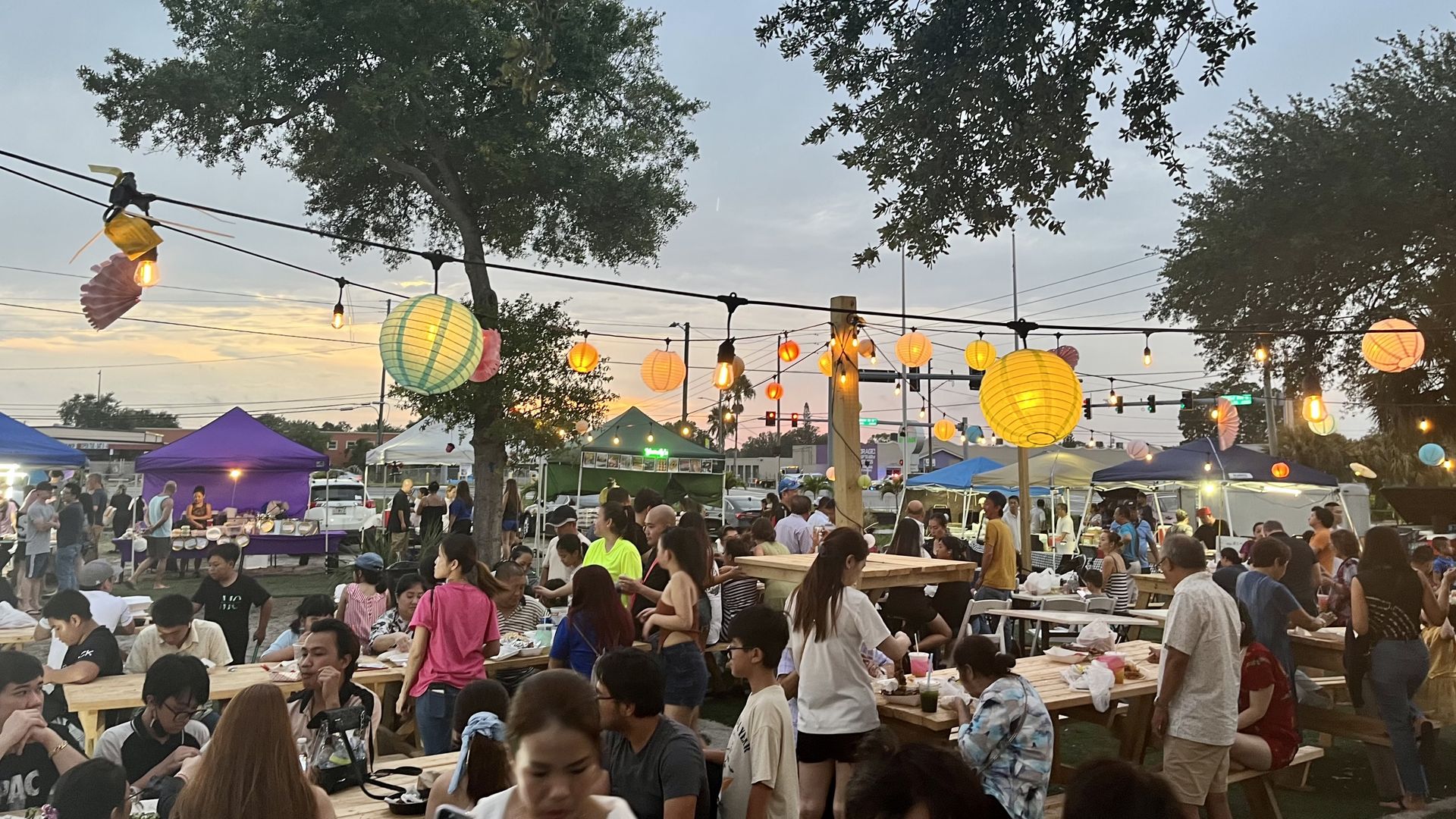 A group of people eating at picnic tables beneath a string of paper lanterns.