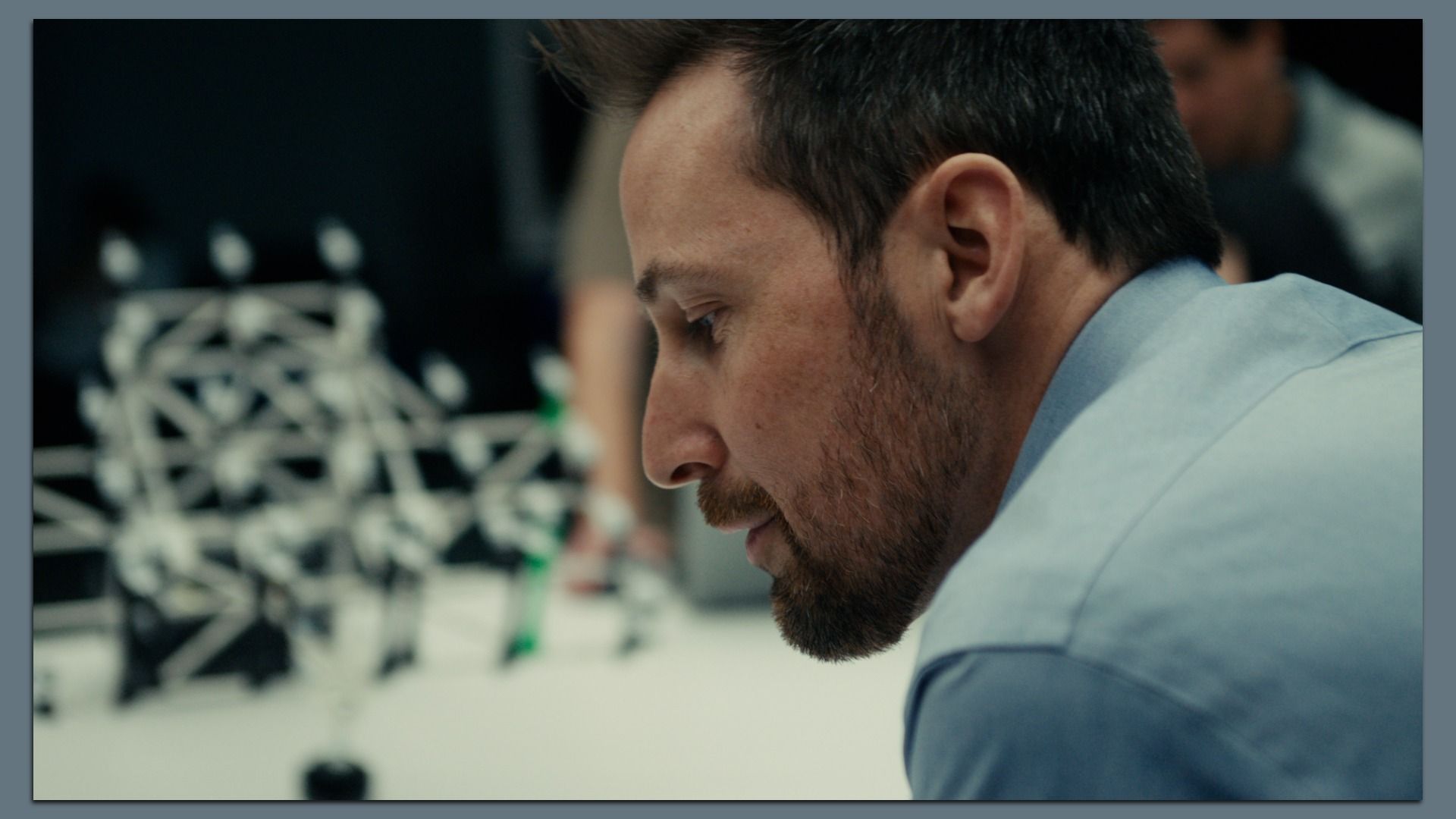 Close-up side profile of Ford Vice President Alan Clarke, with short dark hair and a beard, wearing a light blue shirt, looking downward. A blurred lab or workshop setup with geometric structures appears in the background.