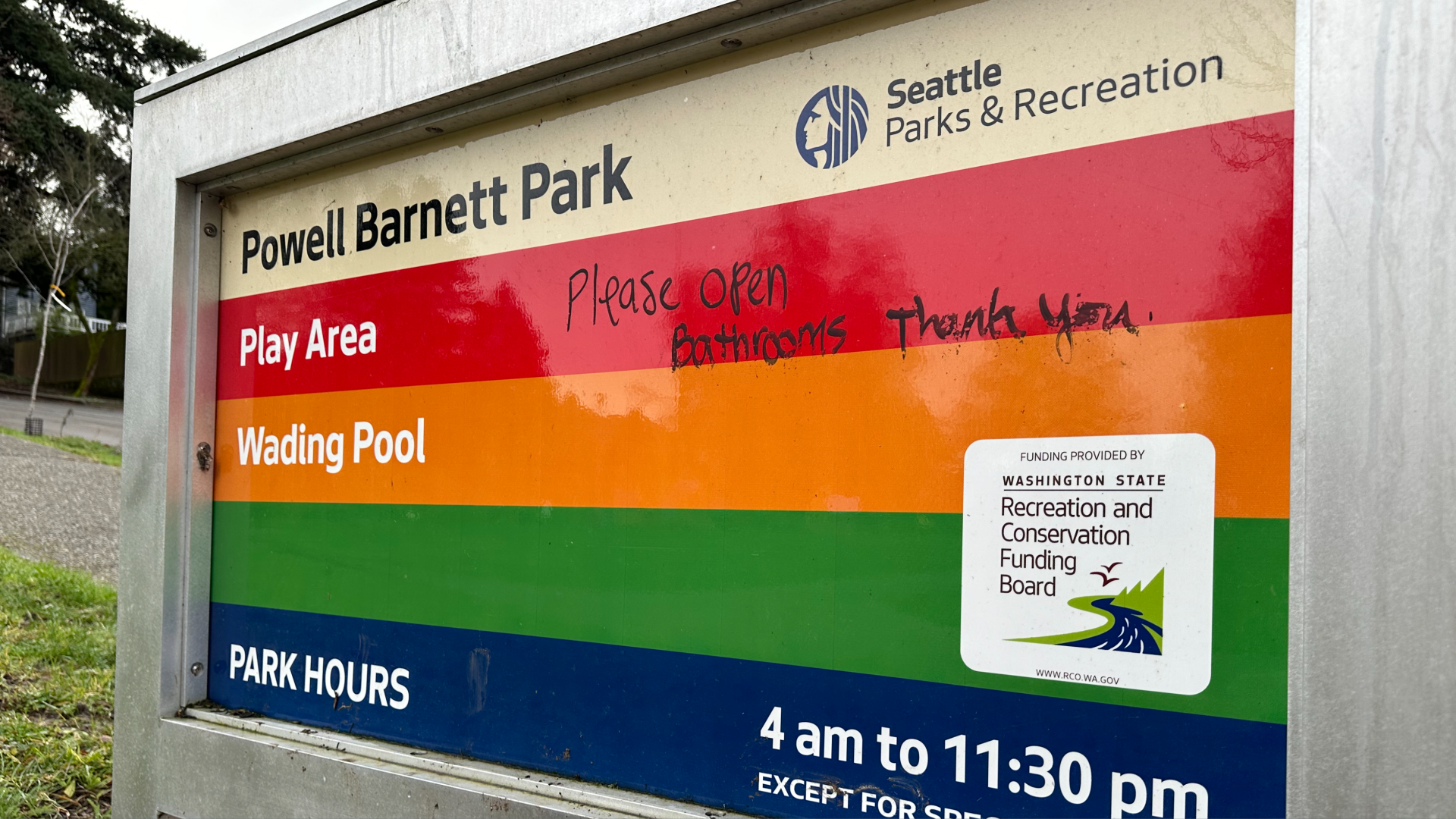 A sign for Seattle's Powell Barnett Park has a message in black ink, handwritten, saying "Please open Bathrooms Thank you" alongside the park hours and listing of play area and wading pool.