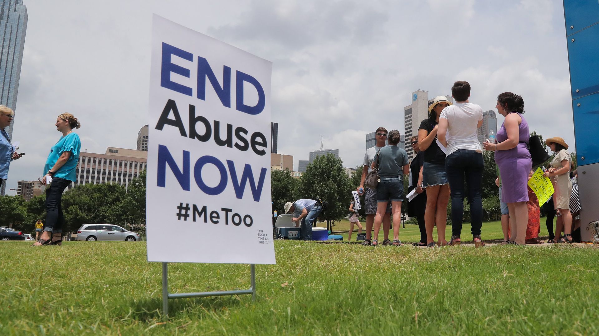 Photo of a sign that says "End Abuse Now #MeToo" embedded in grass with people standing around