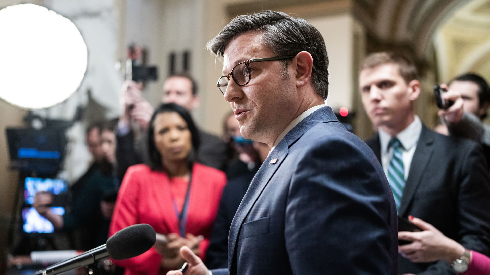 House Speaker Mike Johnson, wearing a blue suit and standing at a microphone, speaks to reporters in the Capitol.