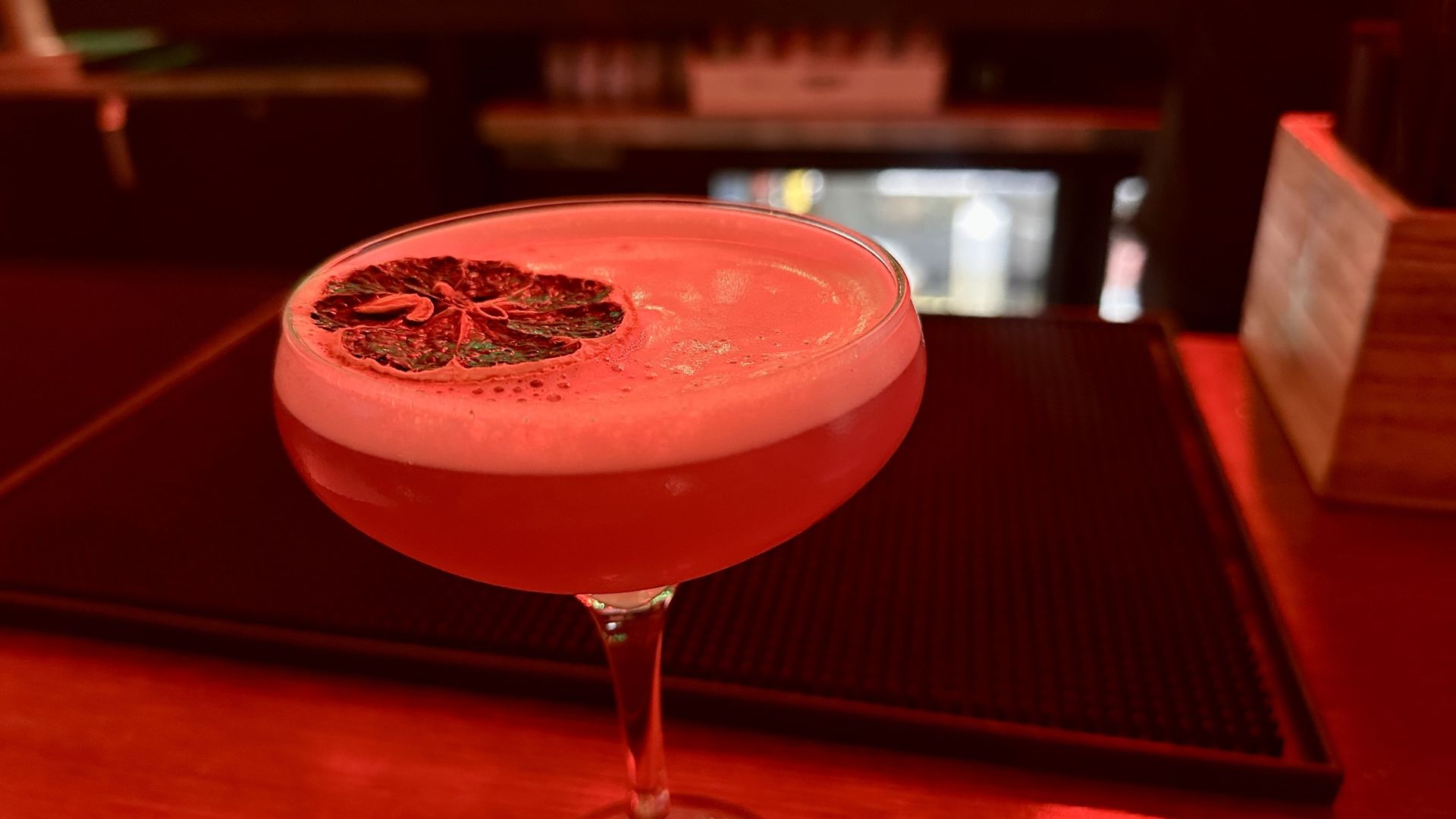 Blue-ish cocktail in a coupe glass with a foam top and a dried citrus slice garnish, set on a bar counter with a soft red light ambiance.