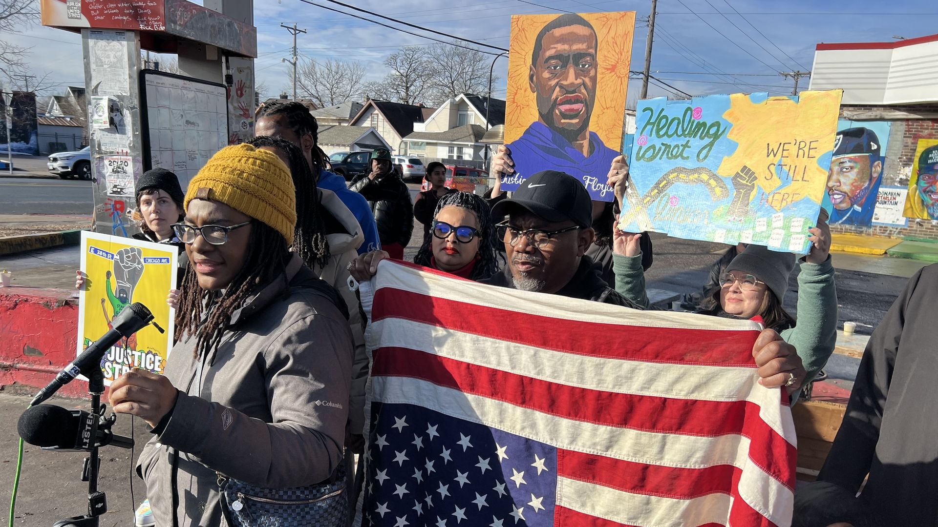 A woman with dark skin in a jacket and yellow hat speaks at a microphone as protestors hold up signs and a painting of George Floyd