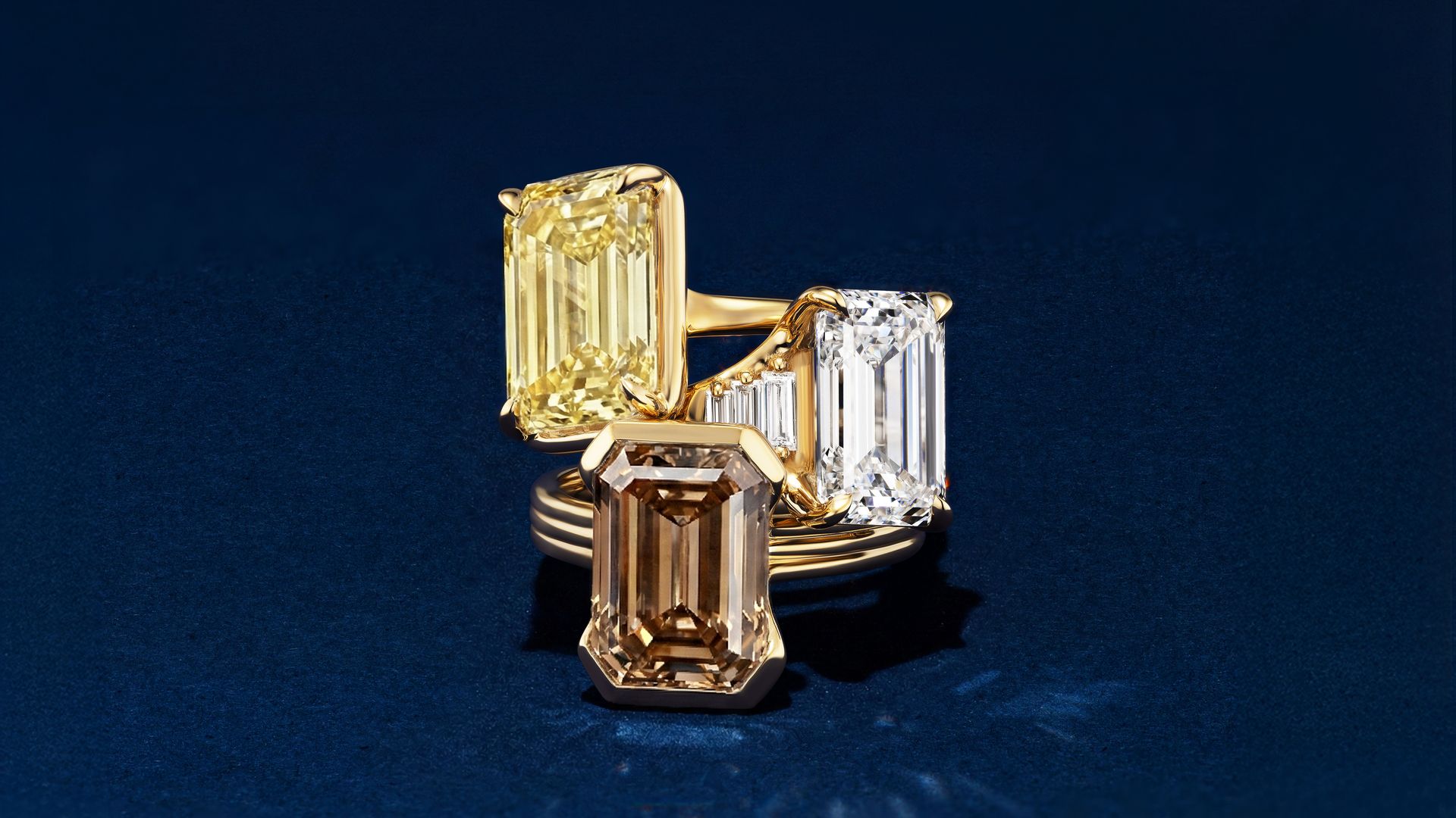 Three gold rings with large emerald-cut diamonds in yellow, clear, and light brown set against a dark blue background.