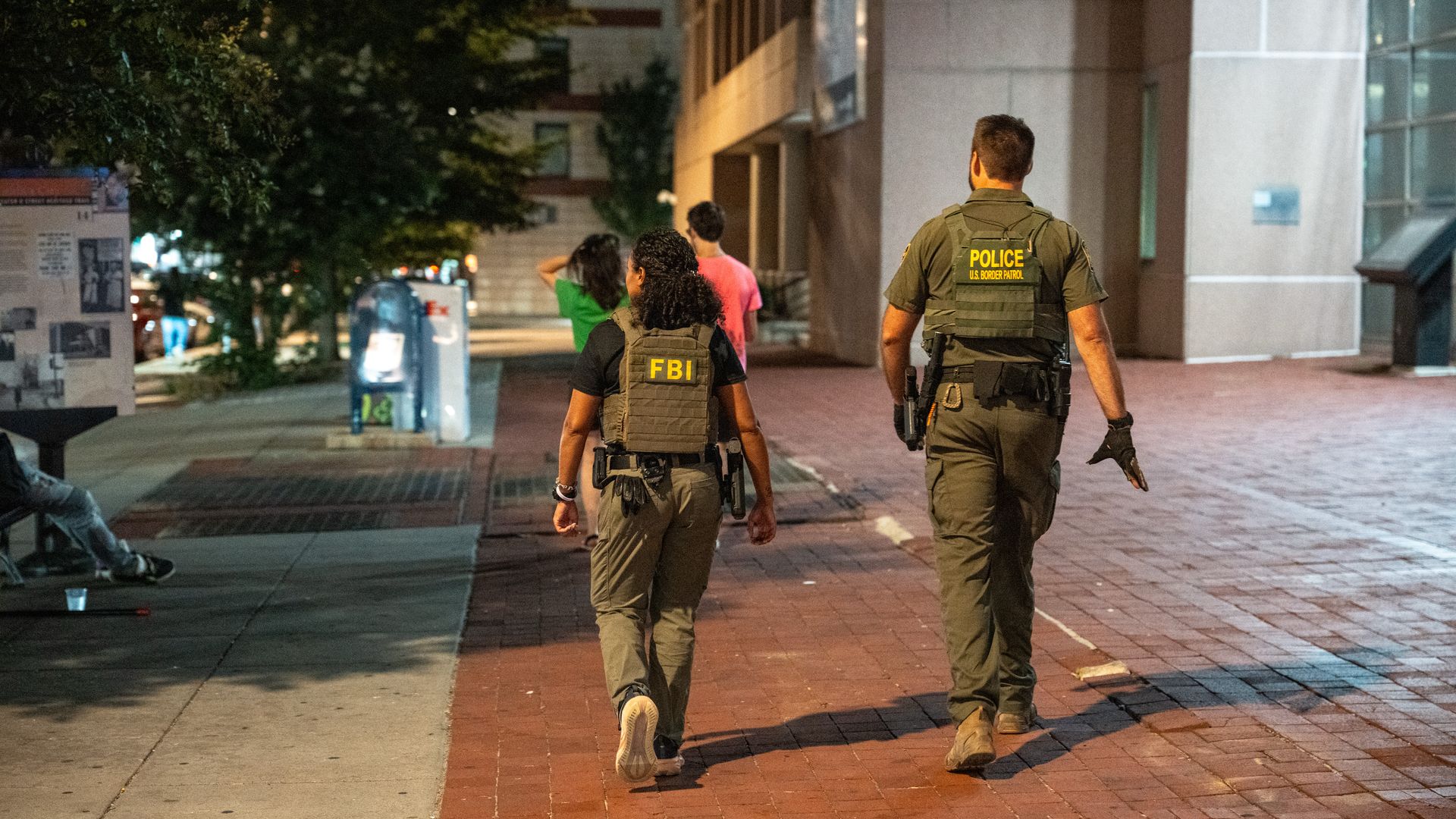 Two law enforcement officers, one with an FBI vest and the other with a U.S. Border Patrol police vest, walk on a brick sidewalk in an urban area at night.