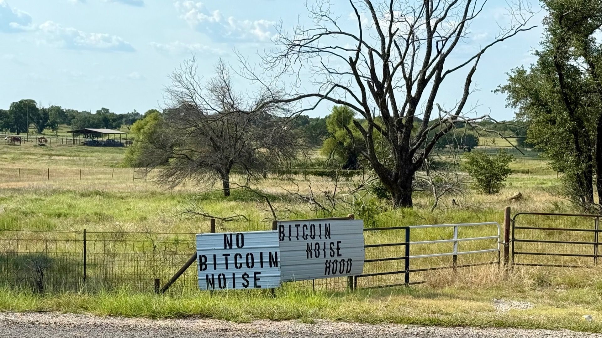 Two white signs on a rural fence read "No Bitcoin Noise" and "Bitcoin Noise No Hood" against a backdrop of grassy fields, leafless trees, and a partly cloudy sky.