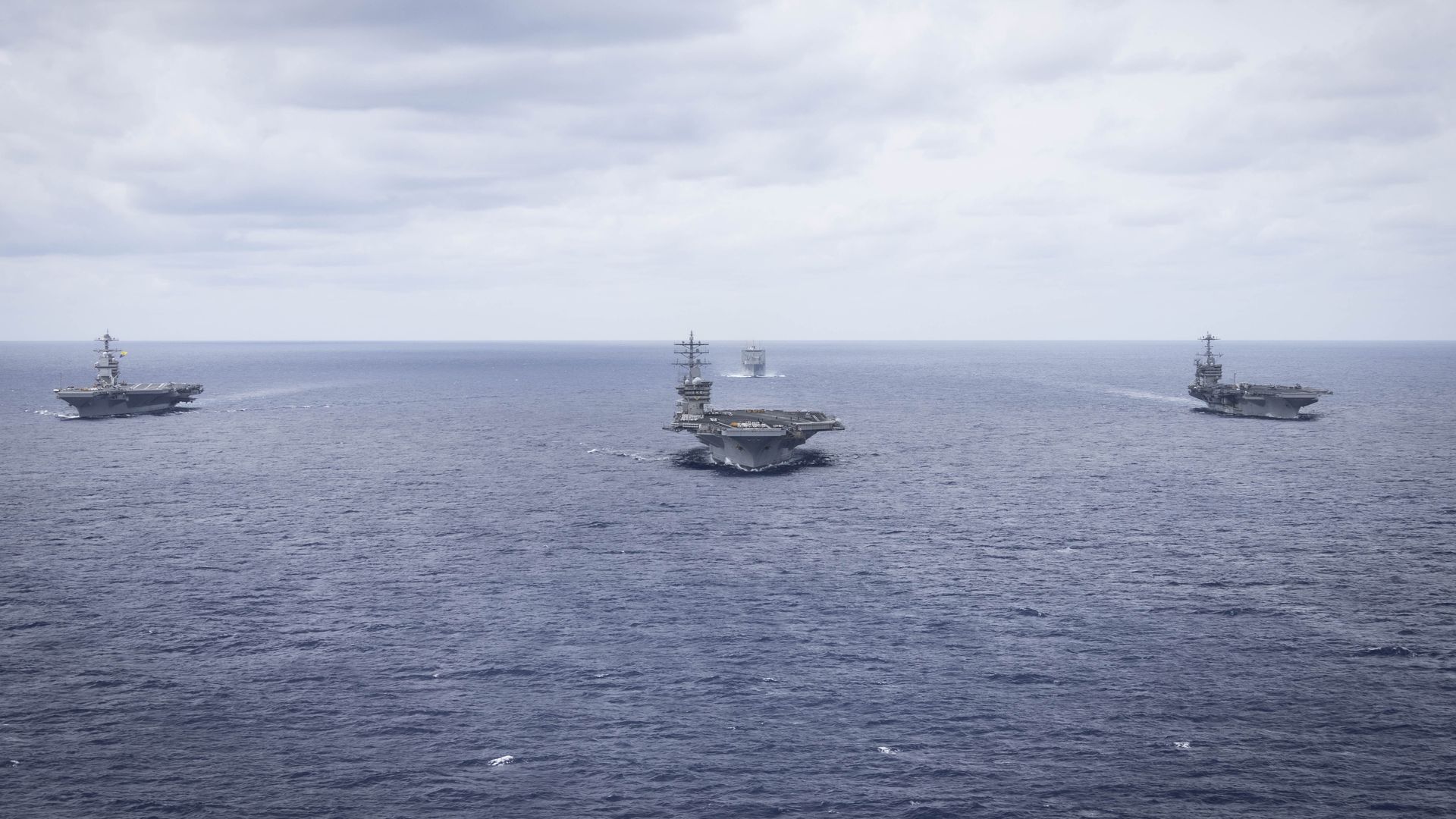 U.S. Navy aircraft carriers glide through the Atlantic Ocean. The water and skies are blue. Clouds are overhead, as well.