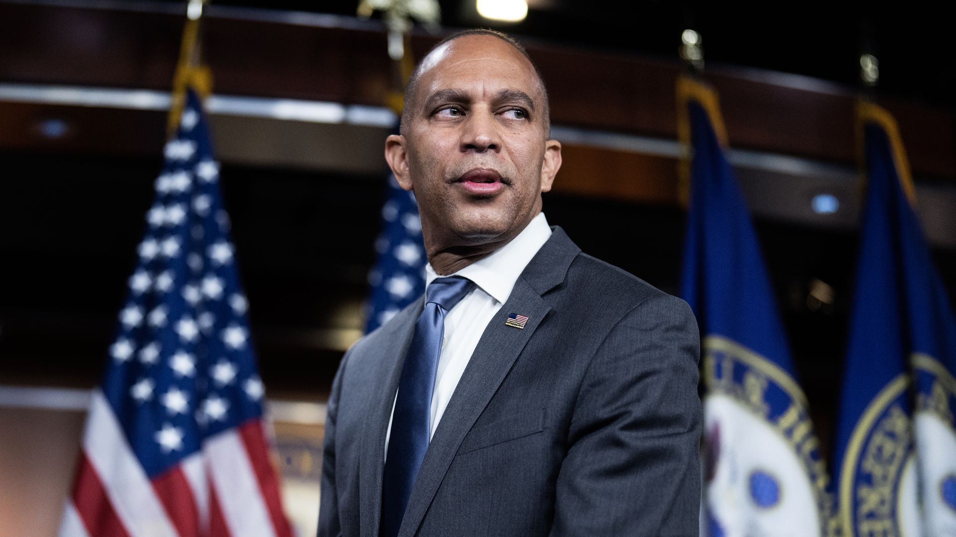 House Minority Leader Hakeem Jeffries, wearing a blue suit and standing in front of flags.