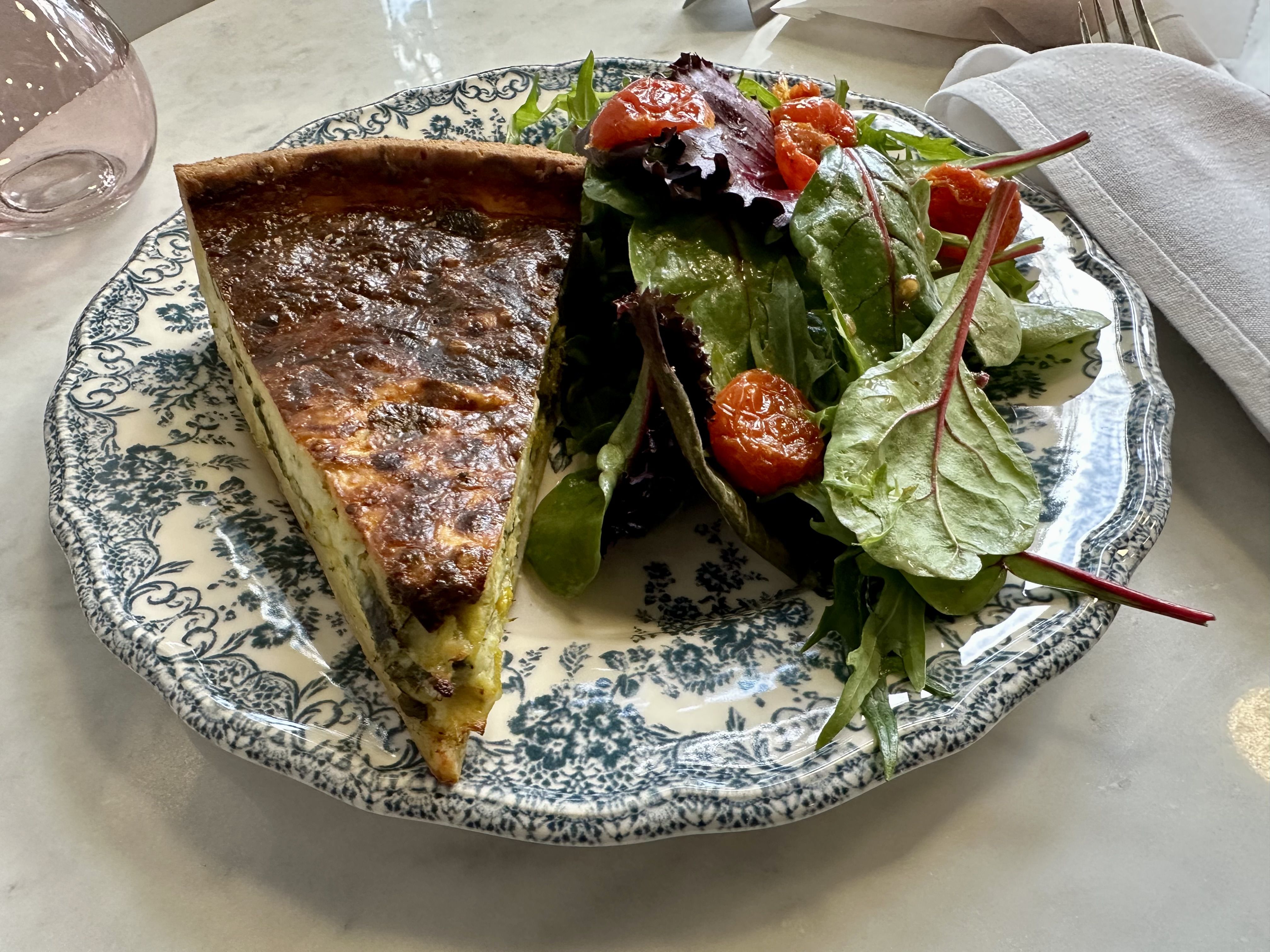 A piece of quiche on a blue and white ceramic plate, with a salad of mixed greens and tomatoes. 