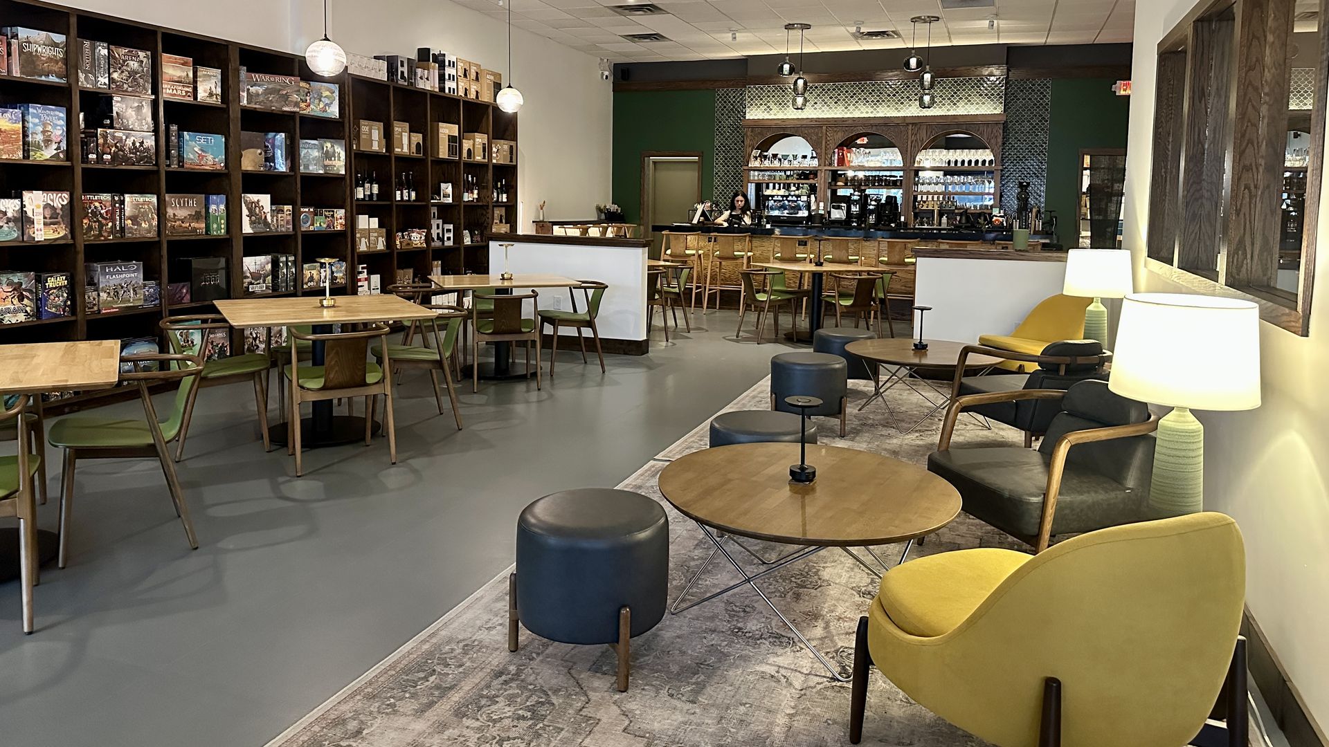 Interior of a cozy cafe with green and yellow chairs, wooden tables, a bar with stools, and shelves filled with board games along the left wall under hanging lights.