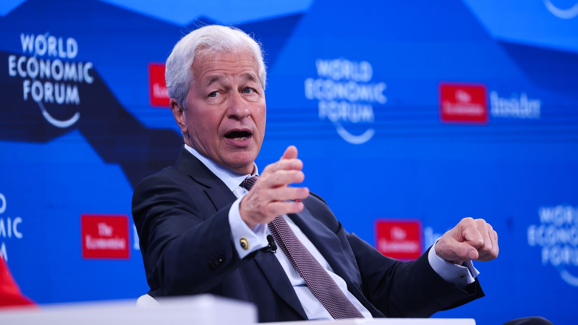 Person with white hair in a dark suit and tie speaking and gesturing with his hand at the World Economic Forum, with blue background featuring event logos and sponsor names.