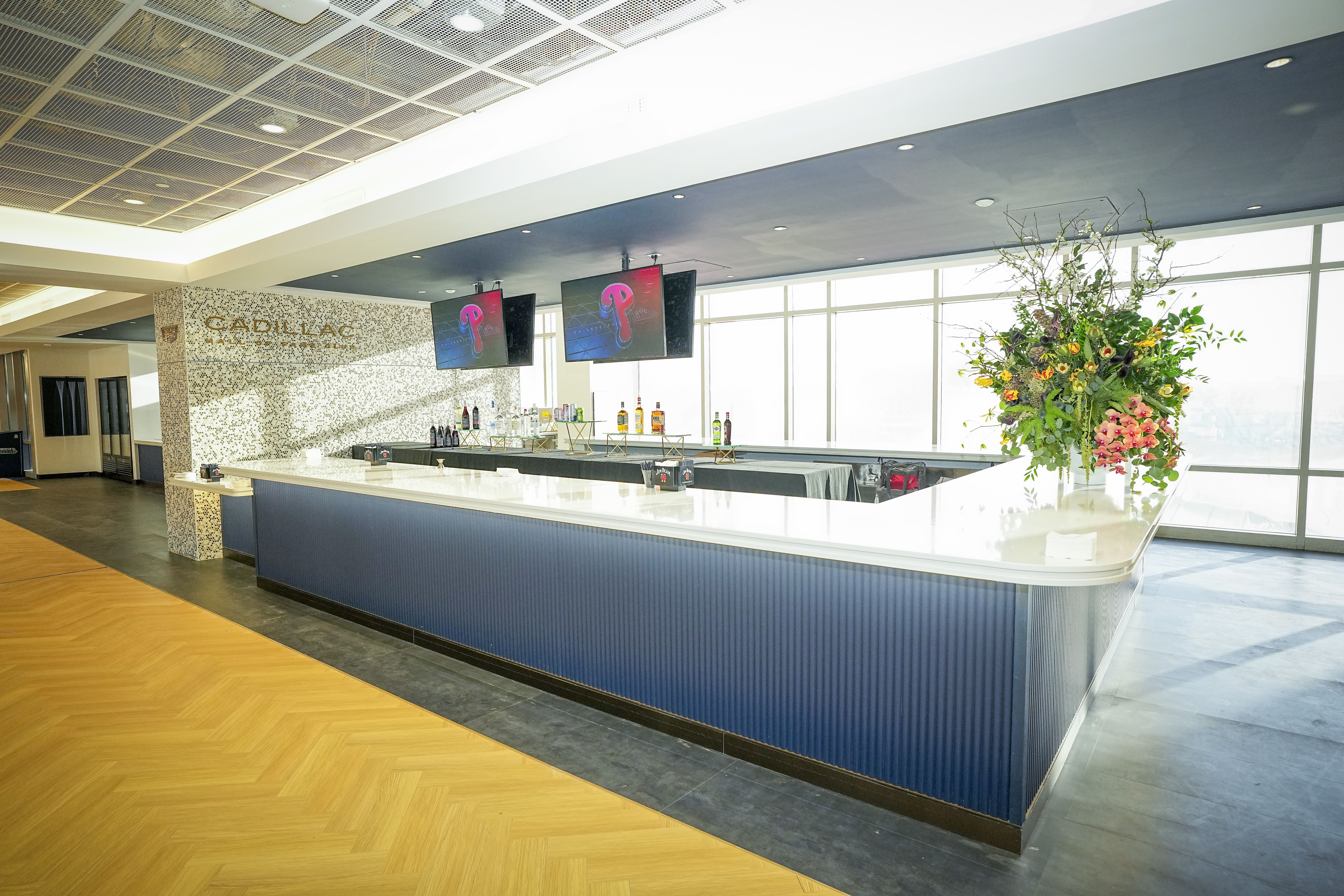 Modern lounge bar with a curved blue counter, a row of bottles and taps behind, two screens showing a pink logo, a tall flower arrangement on the right, and a mosaic Cadillac wall by large windows.